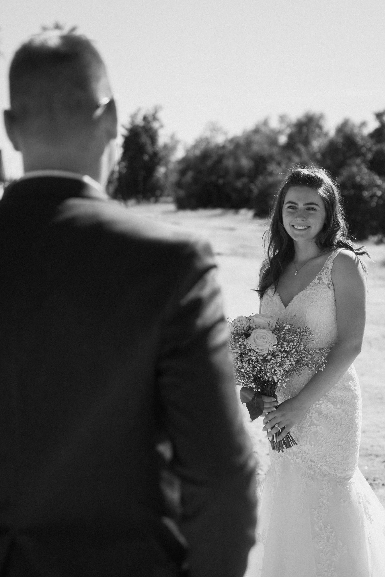 Smiling bride in lace wedding dress holds bouquet while facing groom outdoors in black and white photo.