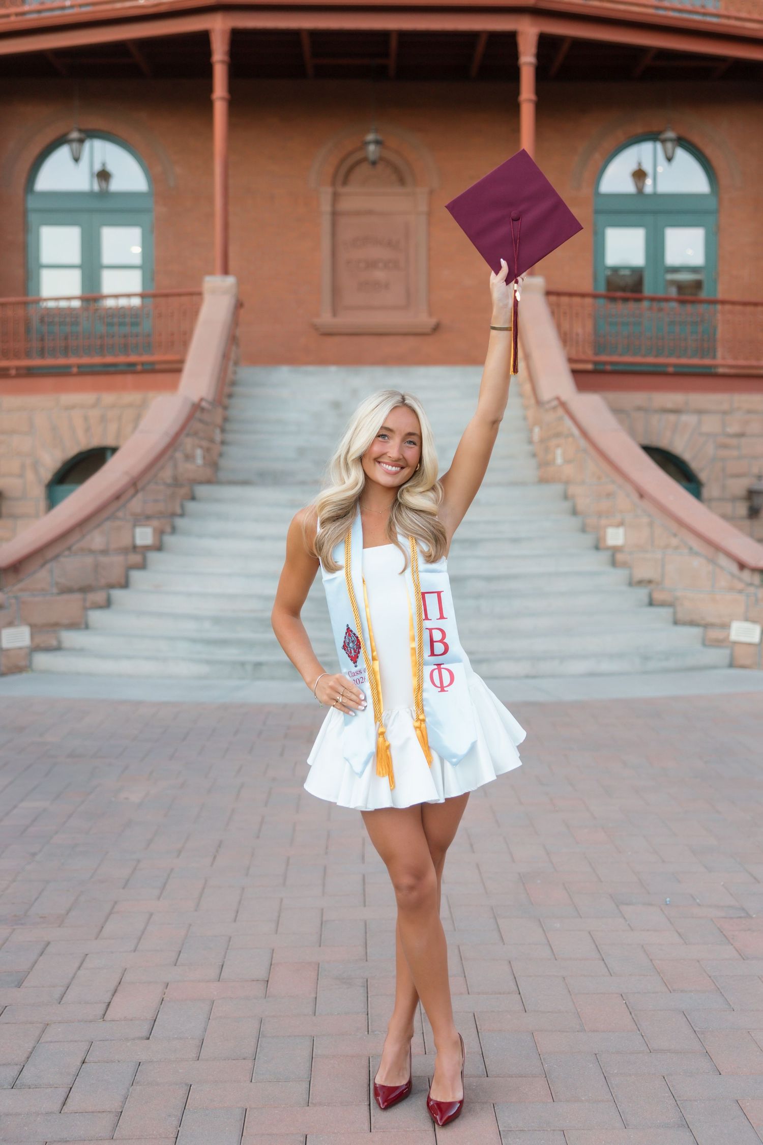 Smiling blonde graduate in white dress and Pi Beta Phi sash raises maroon cap outdoors on brick plaza.