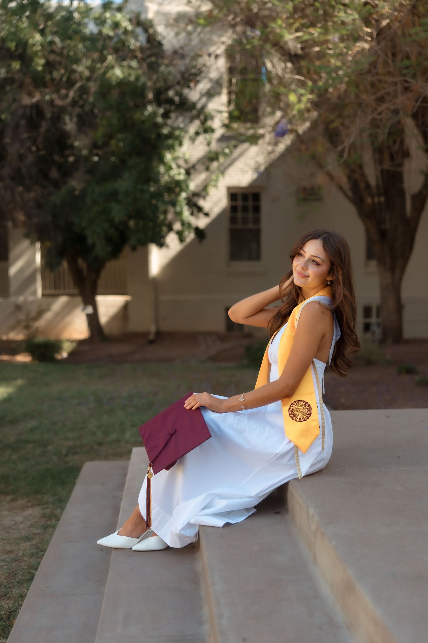Young woman in white dress and graduation stole sits on steps holding maroon cap outdoors.