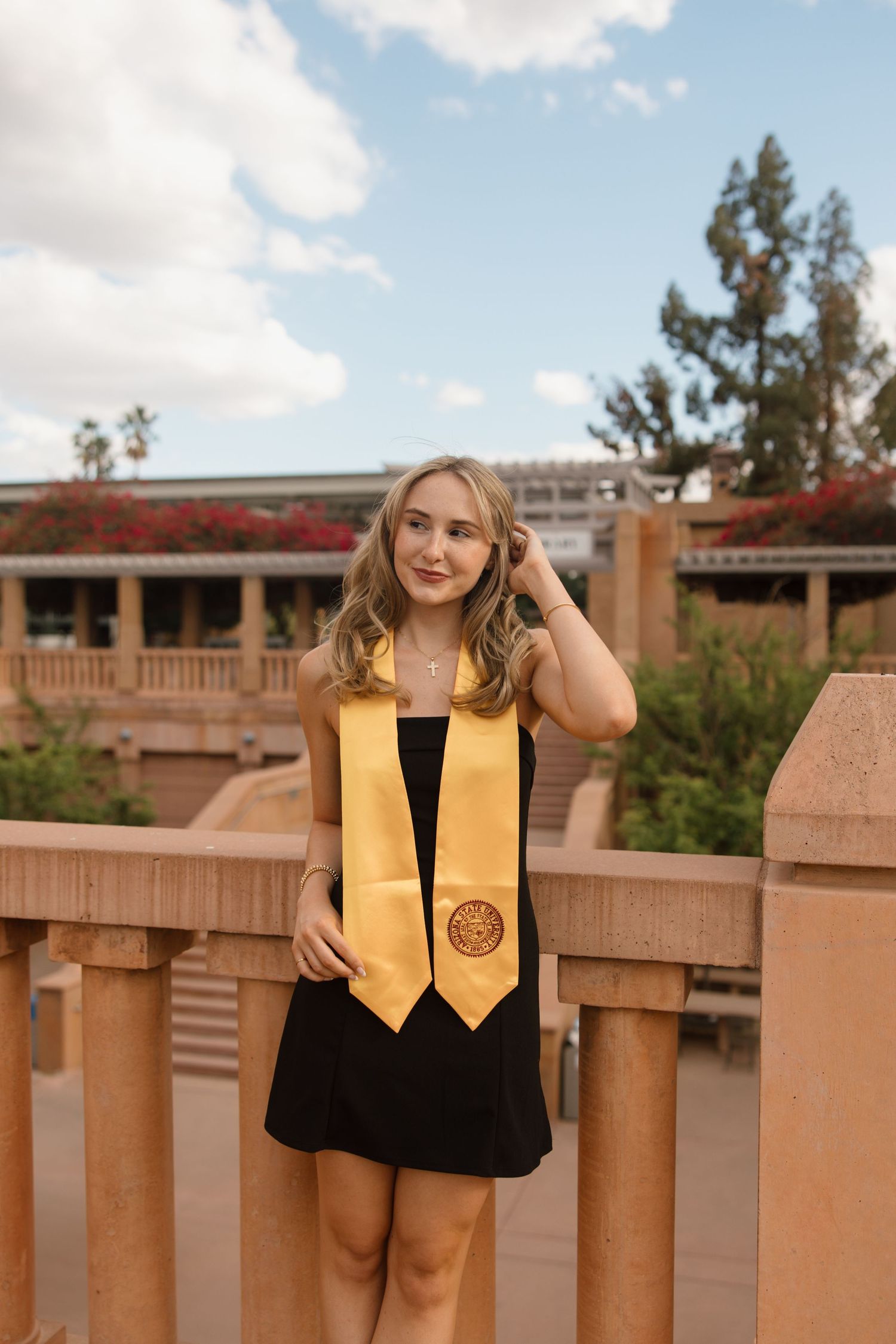 Young woman in black dress and gold graduation stole smiling outdoors at university campus on a cloudy day.