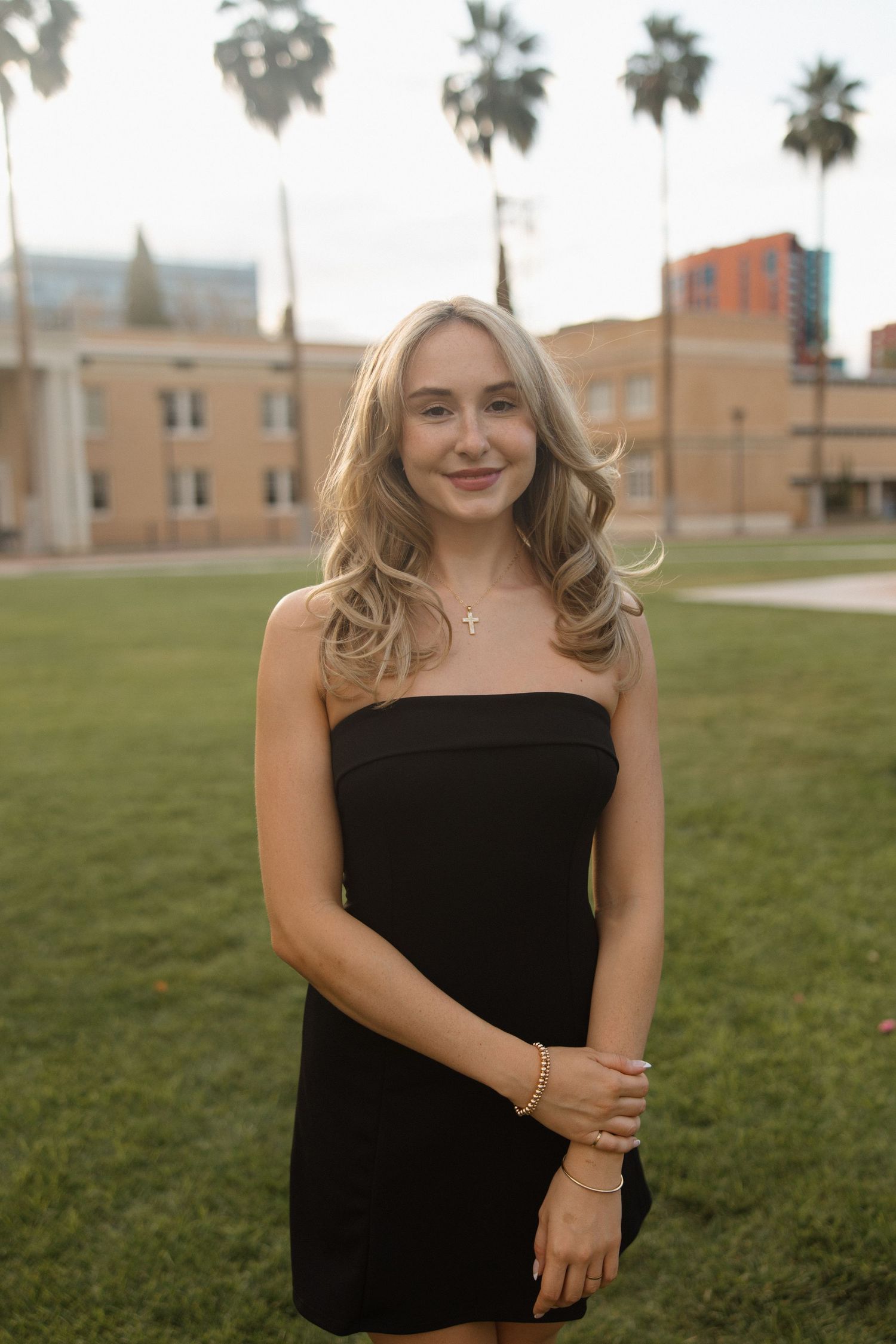 Young blonde woman in black strapless dress smiling outdoors on green lawn with palm trees and buildings in background.