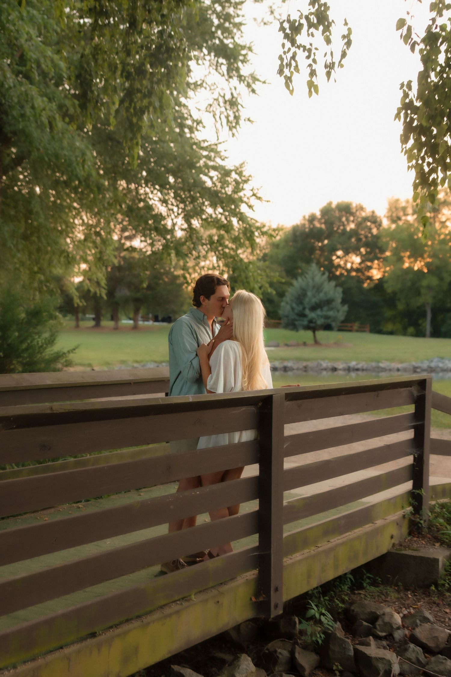 A couple shares a tender kiss on a wooden bridge surrounded by green trees at golden hour.