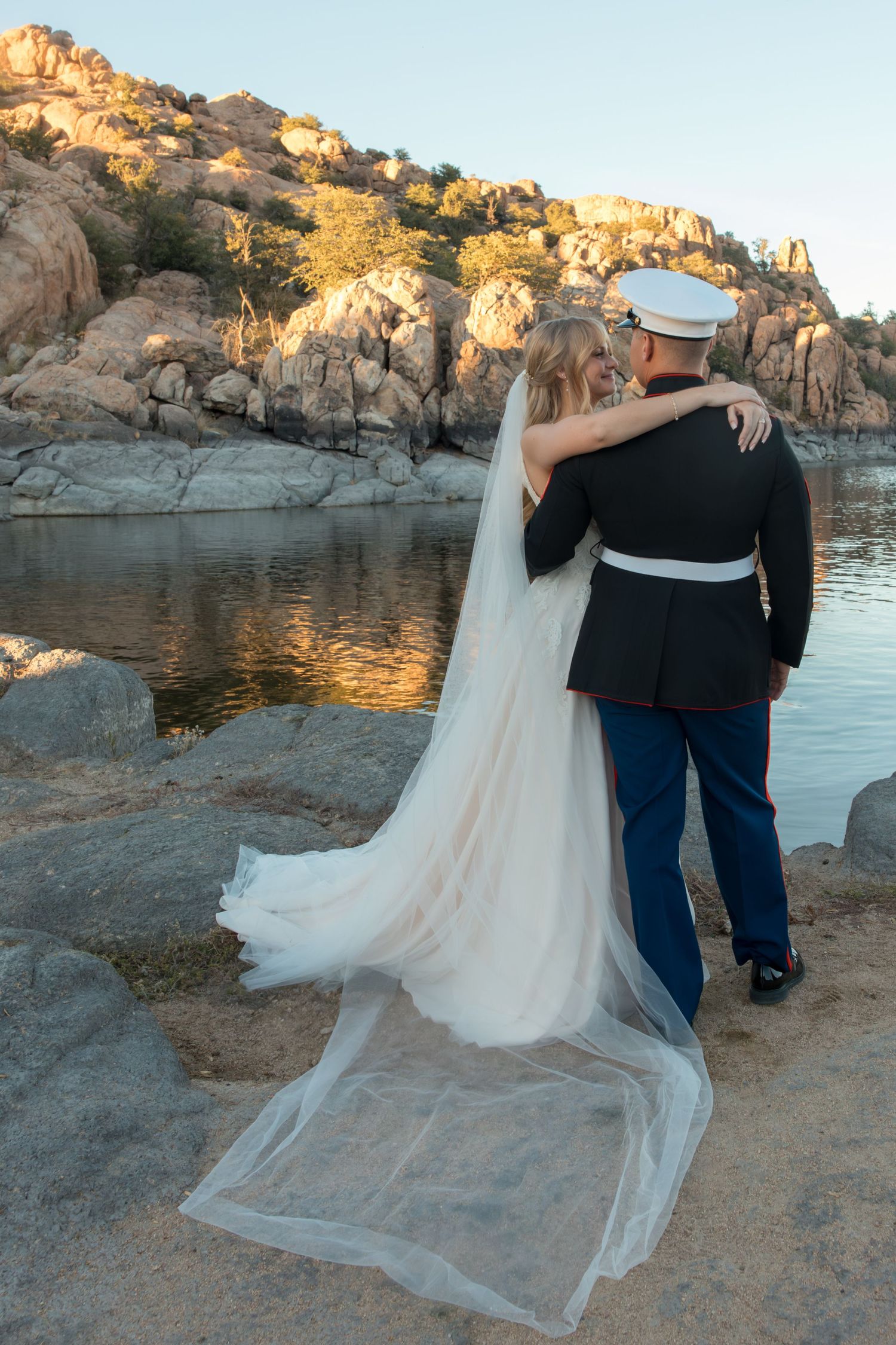 Marine groom in dress blues and bride with cathedral veil stand embracing by a rocky desert lake.