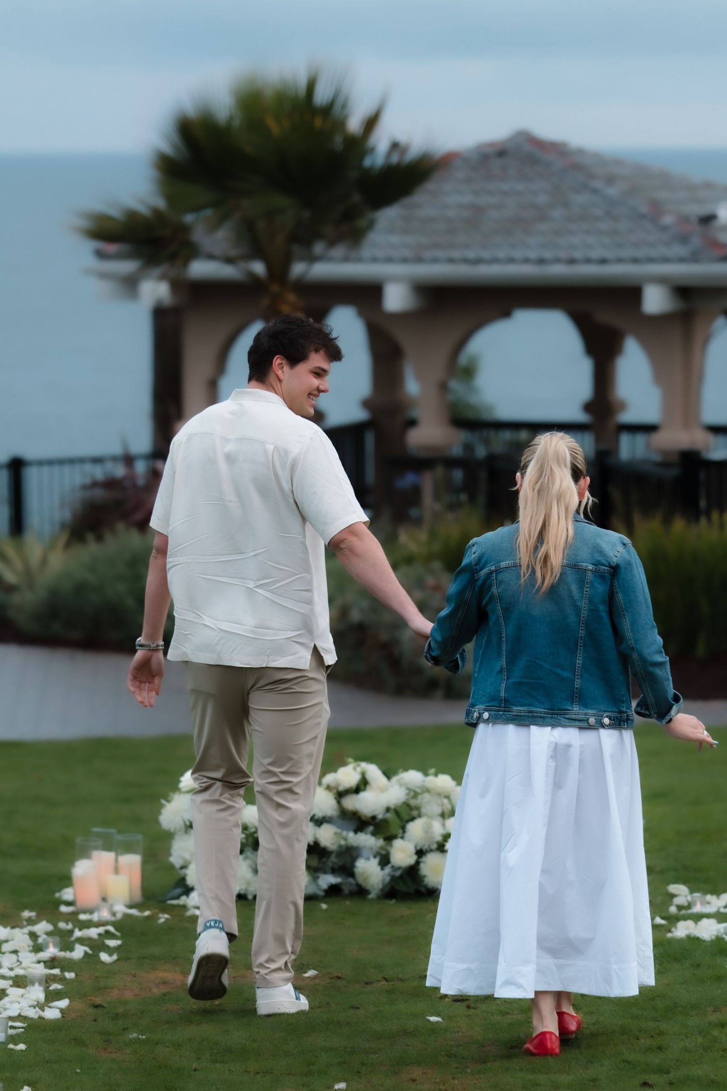 Couple holding hands walking toward ocean view gazebo surrounded by white flowers and candles during romantic proposal setup.