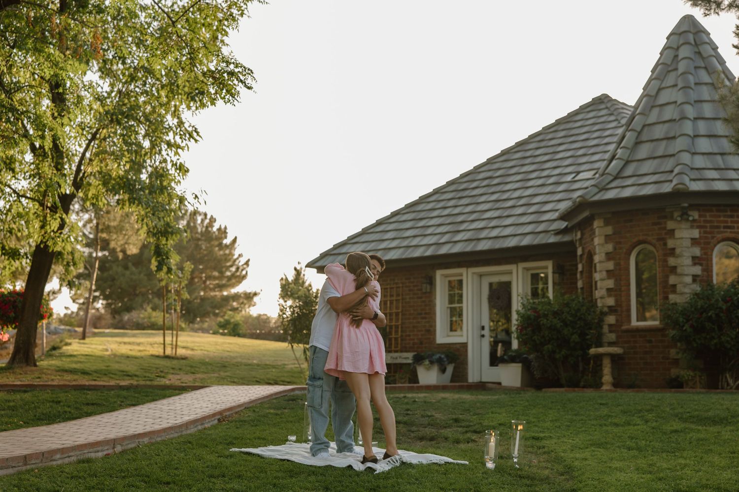 Couple embracing on a picnic blanket in a lush green yard beside a charming brick cottage at sunset.