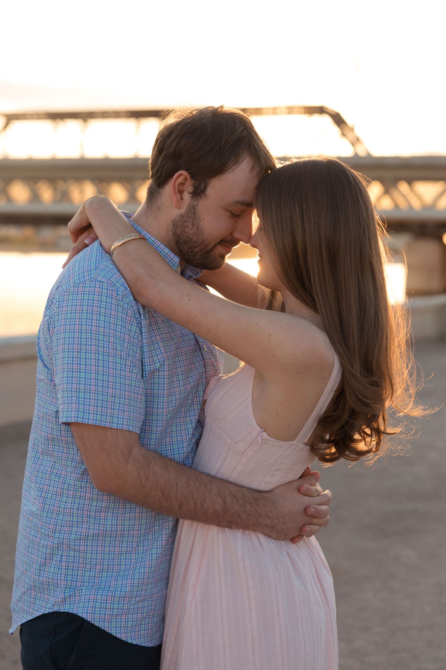 Intimate couple touching foreheads in tender embrace on beach at warm sunset by bridge.