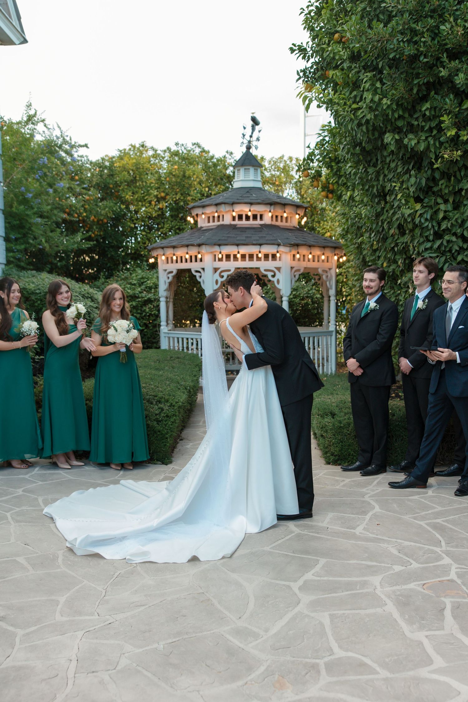 Newlyweds kiss at outdoor gazebo ceremony surrounded by wedding party in emerald green and black attire.