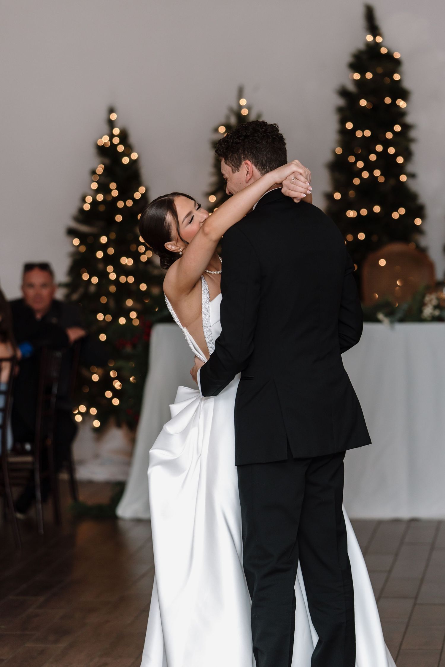 Bride and groom sharing their first dance embracing on the dance floor with glowing Christmas trees behind them.