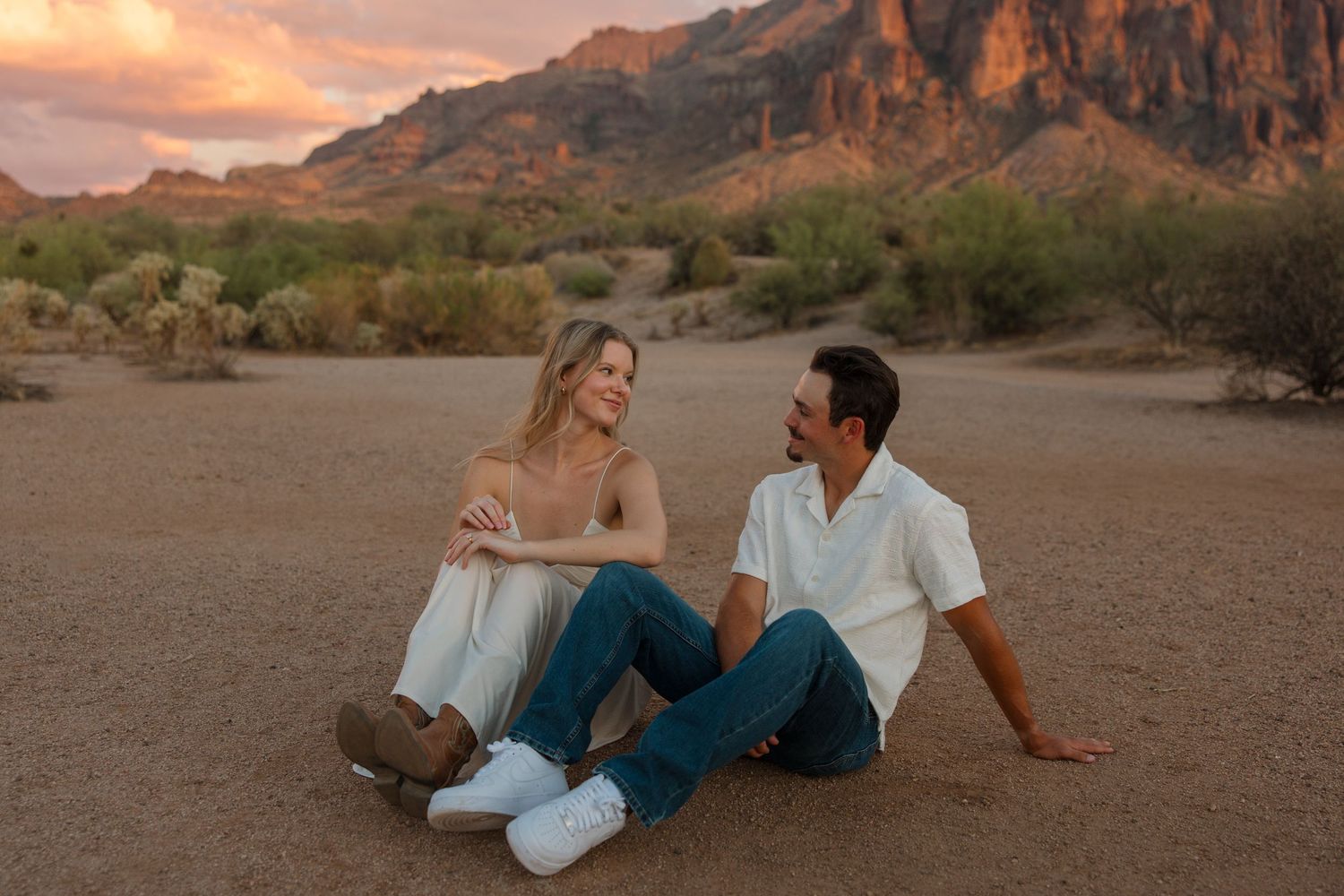 Couple sitting together on desert ground at sunset with dramatic red rock mountains in the background.