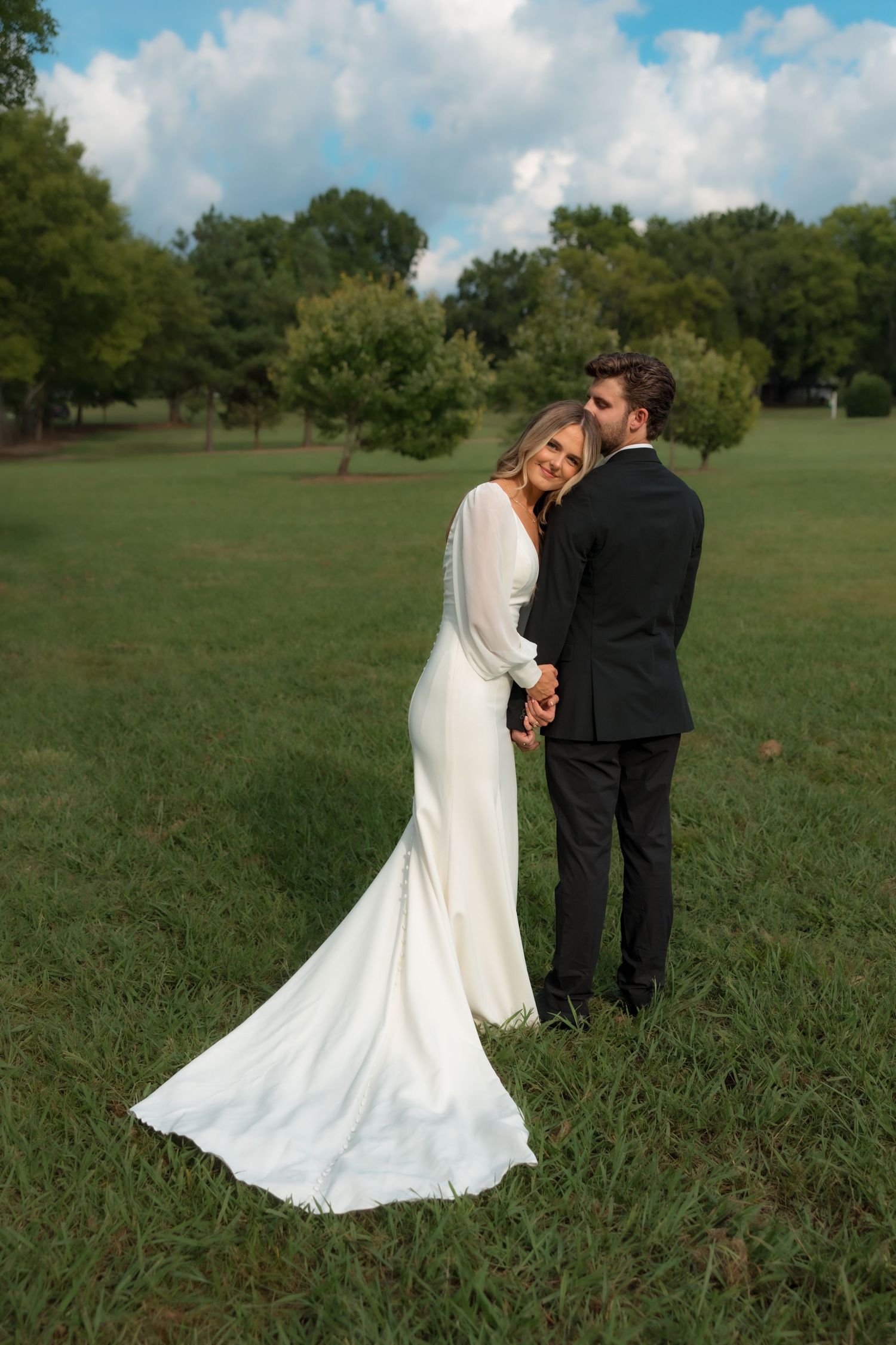 Elegant bride in long-sleeve white gown holds hands with groom in black suit outdoors.