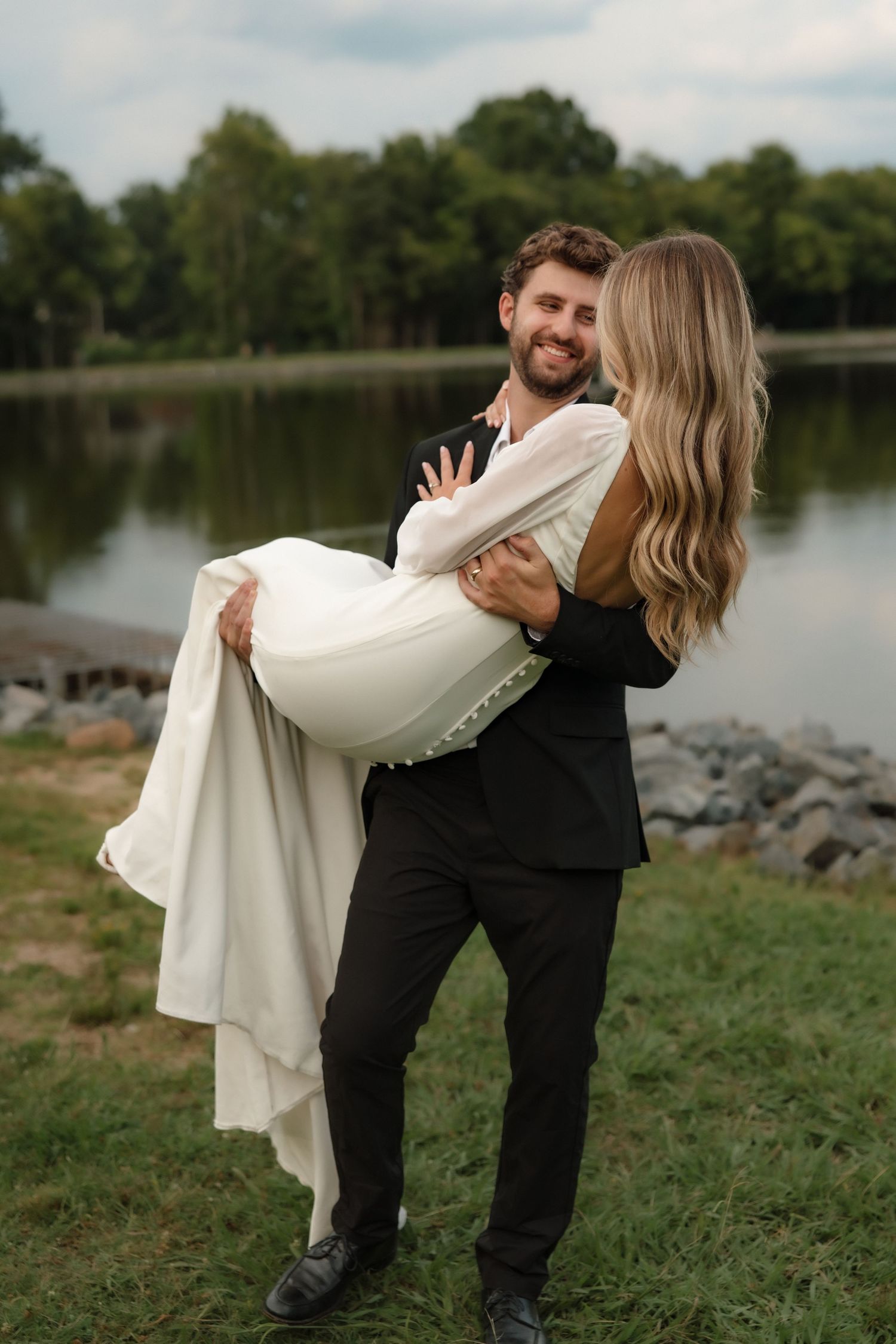 Groom smiles while carrying bride in white wedding dress outdoors near a peaceful lakeside.