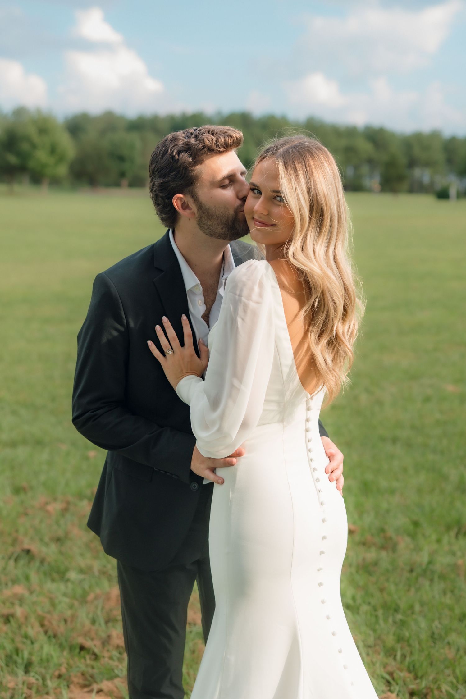 Groom in black suit kissing bride in elegant white backless gown outdoors in green field.