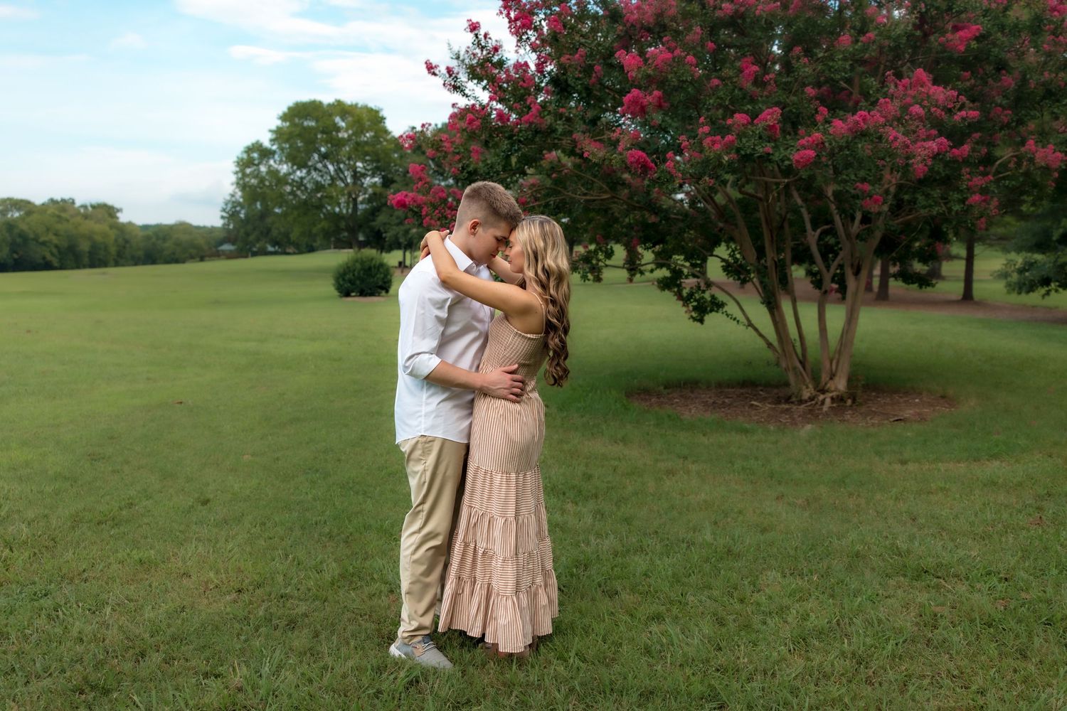 Couple embracing on green lawn near blooming pink crepe myrtle tree during outdoor engagement photo session.
