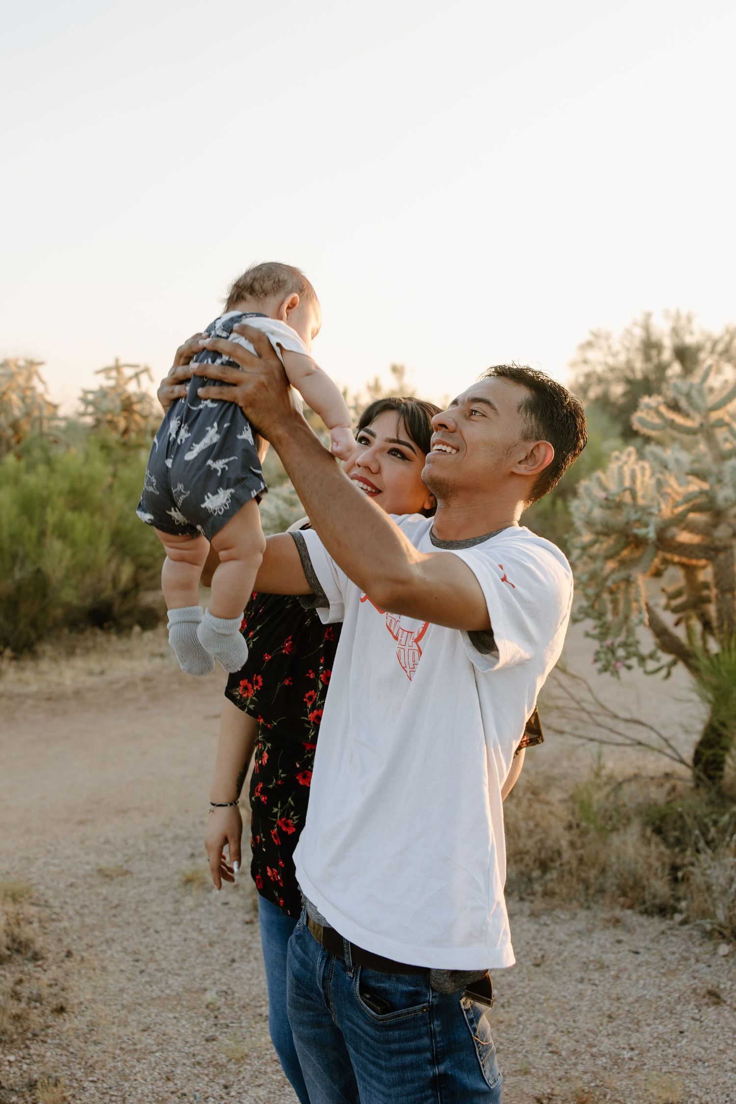 A young couple joyfully lifts their baby in a desert landscape with cacti at golden hour.