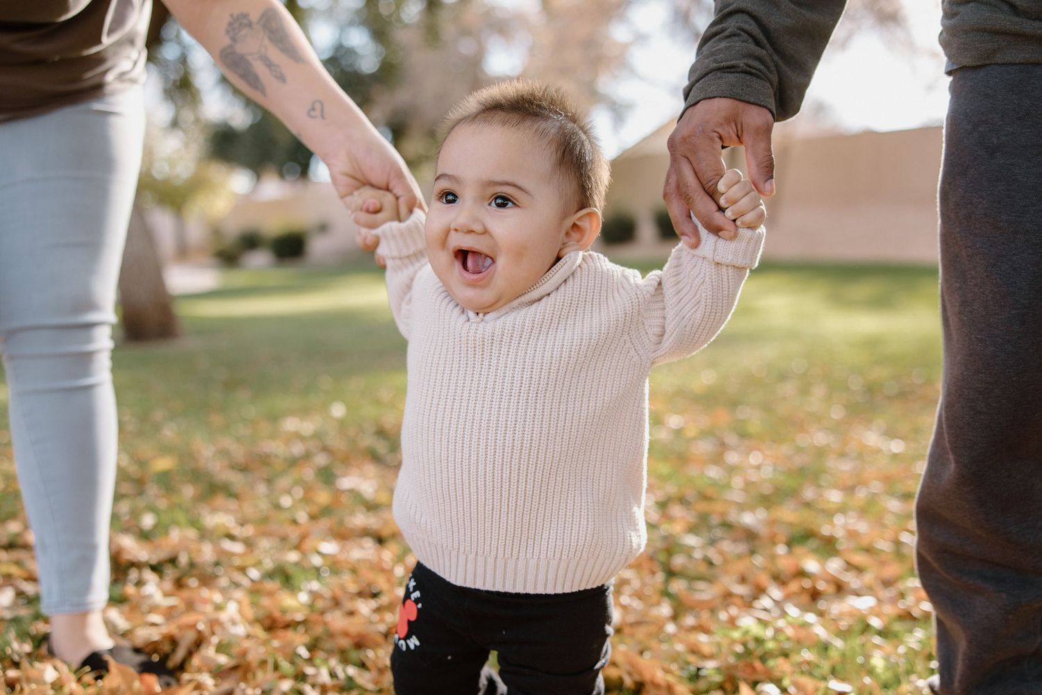 Happy baby in pink sweater holds parents hands while learning to walk outdoors among autumn leaves in a park.
