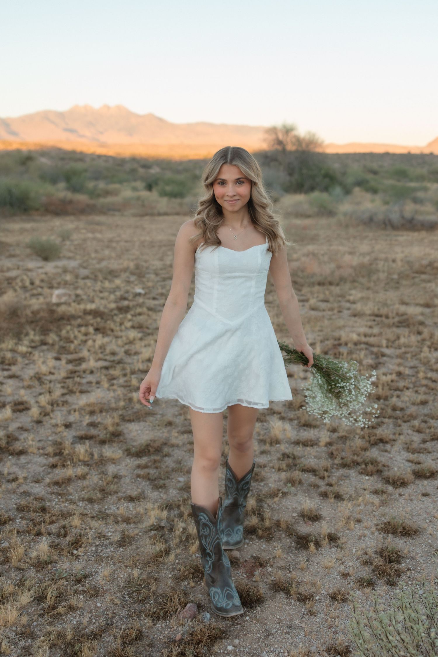 Young woman in white mini dress and cowboy boots holds wildflowers in desert landscape at golden hour.