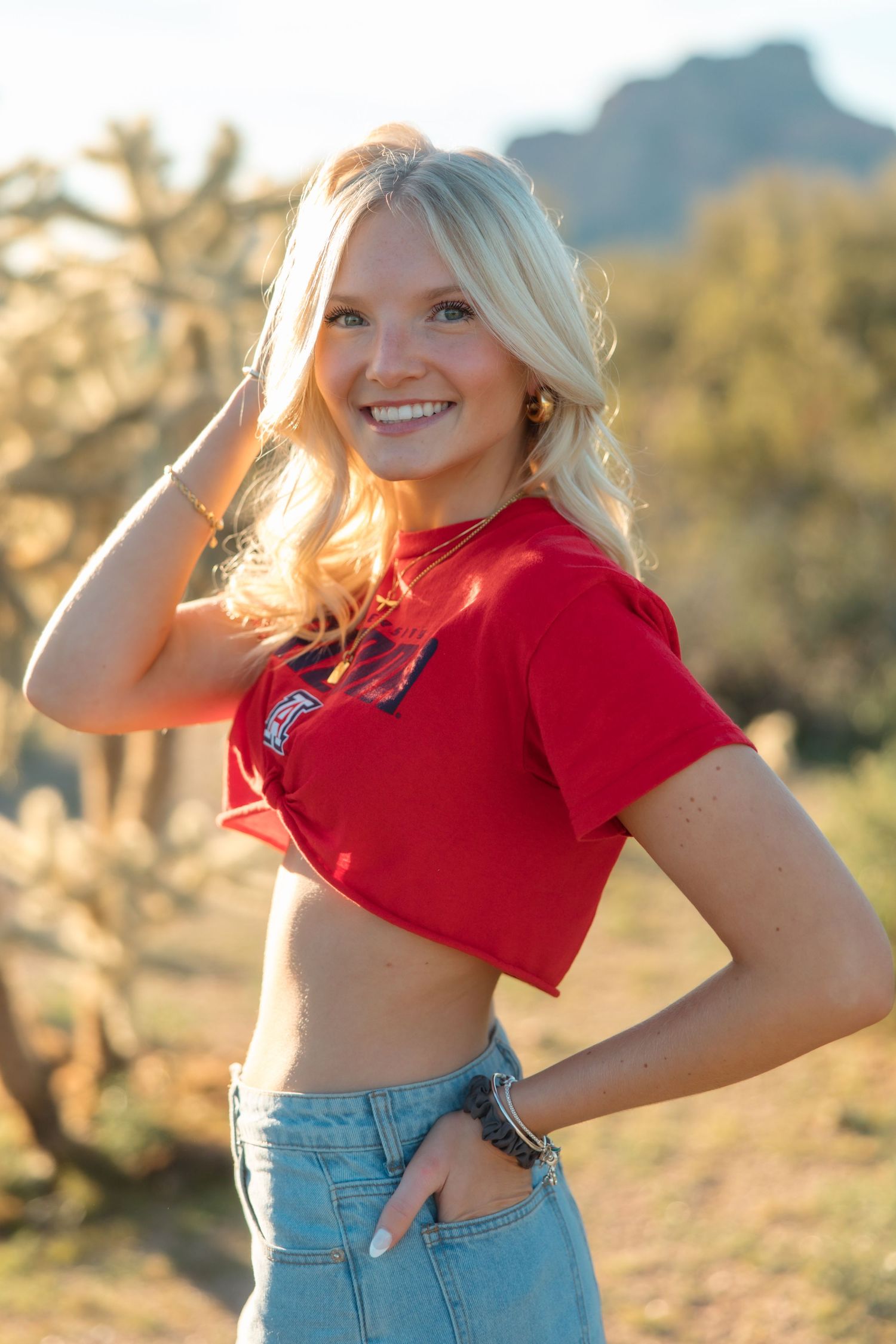 Smiling blonde woman in red crop top and jeans poses outdoors in desert landscape with cacti and mountains.