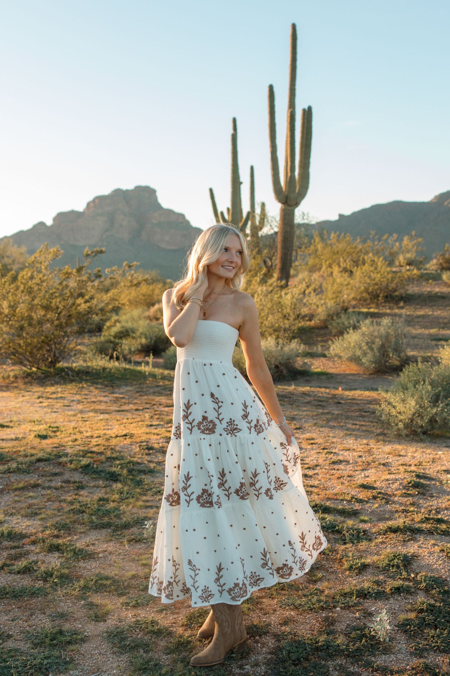 Smiling blonde woman in white floral embroidered strapless midi dress and tan boots in Arizona desert with saguaro cactus.