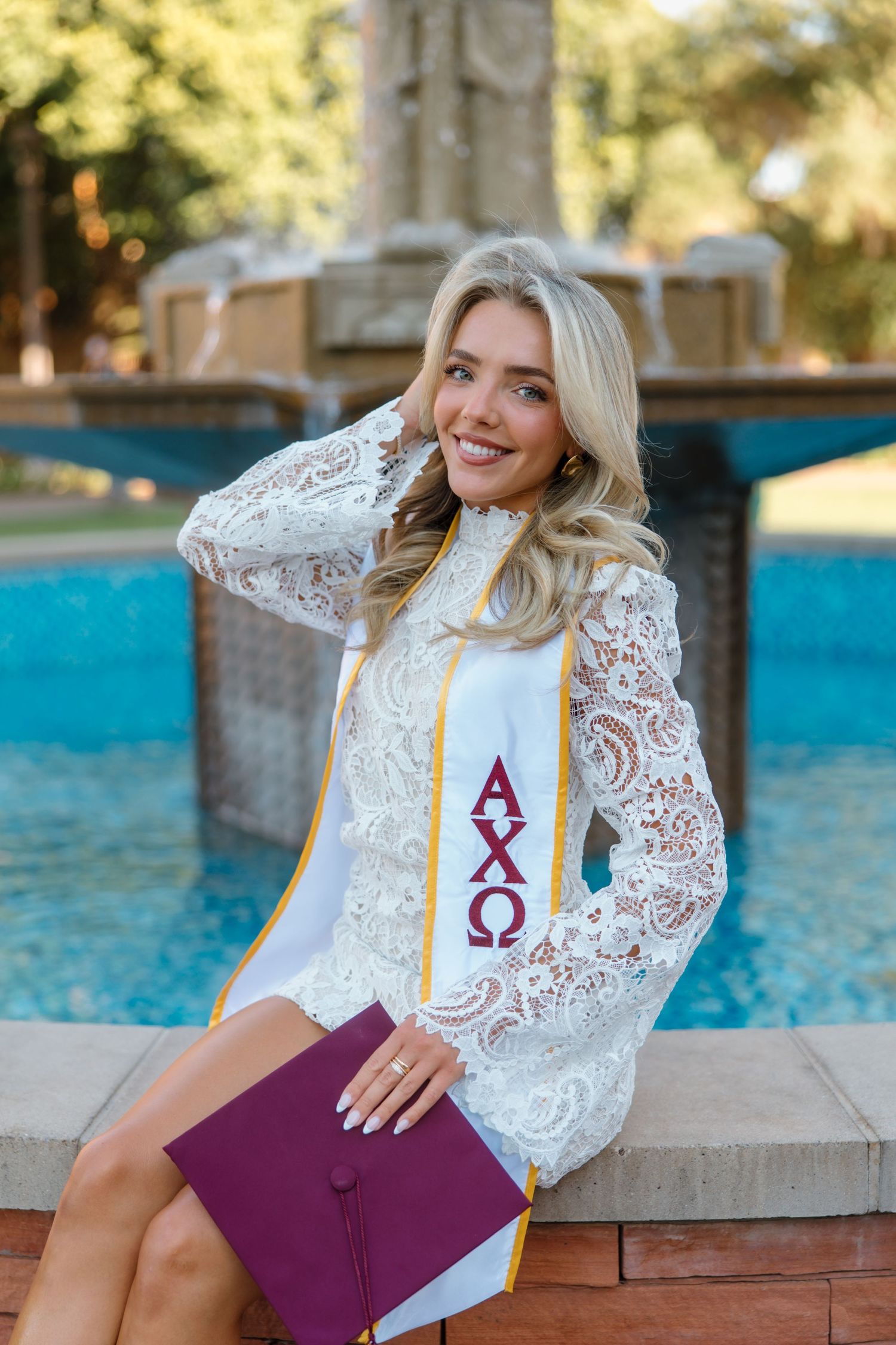 Smiling blonde graduate in white lace dress wearing Alpha Chi Omega stole, holding maroon diploma by fountain.