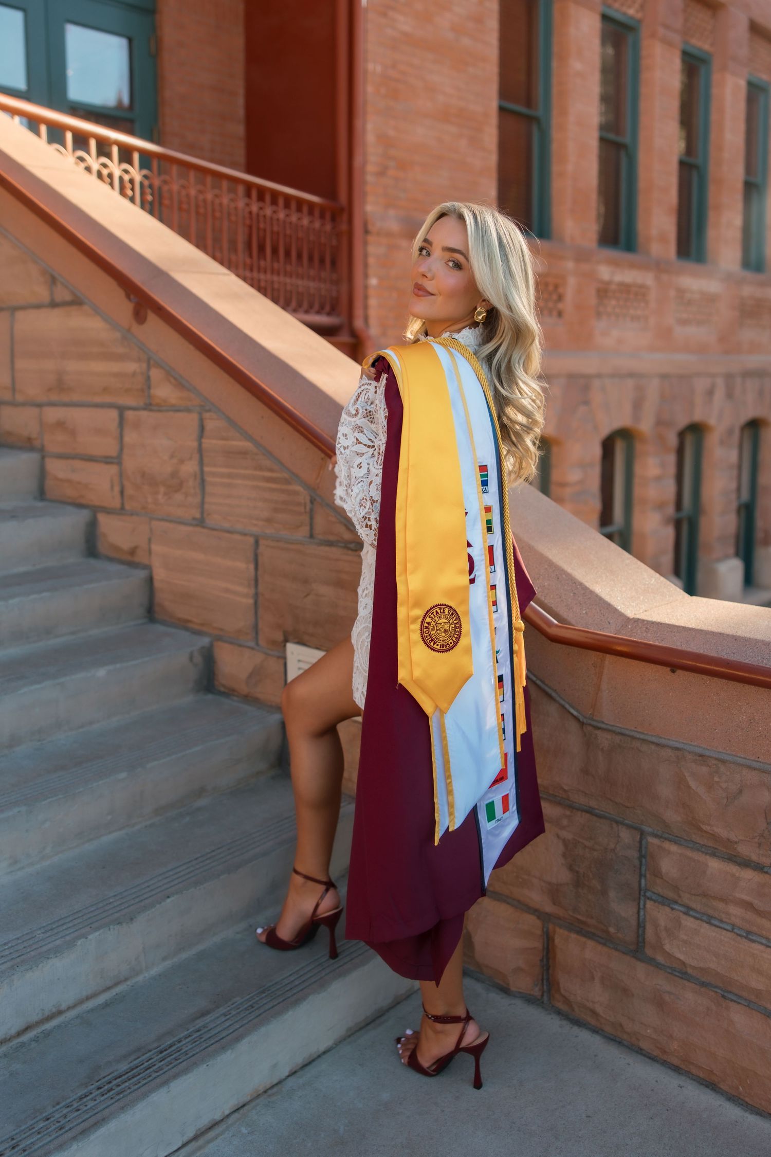 Blonde female graduate poses on brick steps wearing maroon and gold graduation regalia and heels.