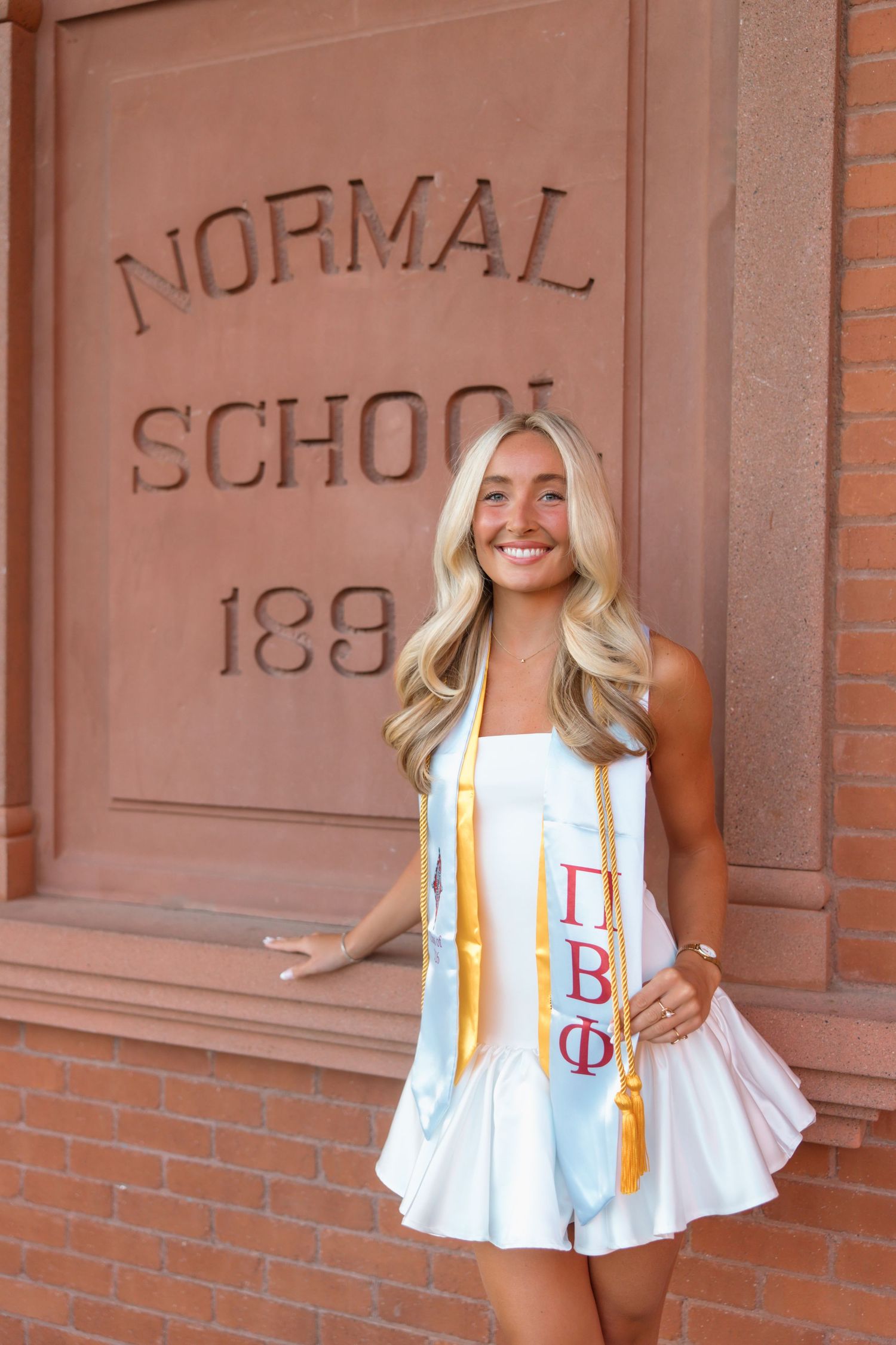 Smiling blonde graduate wearing white dress and Pi Beta Phi stole poses by Normal School 1889 brick building.