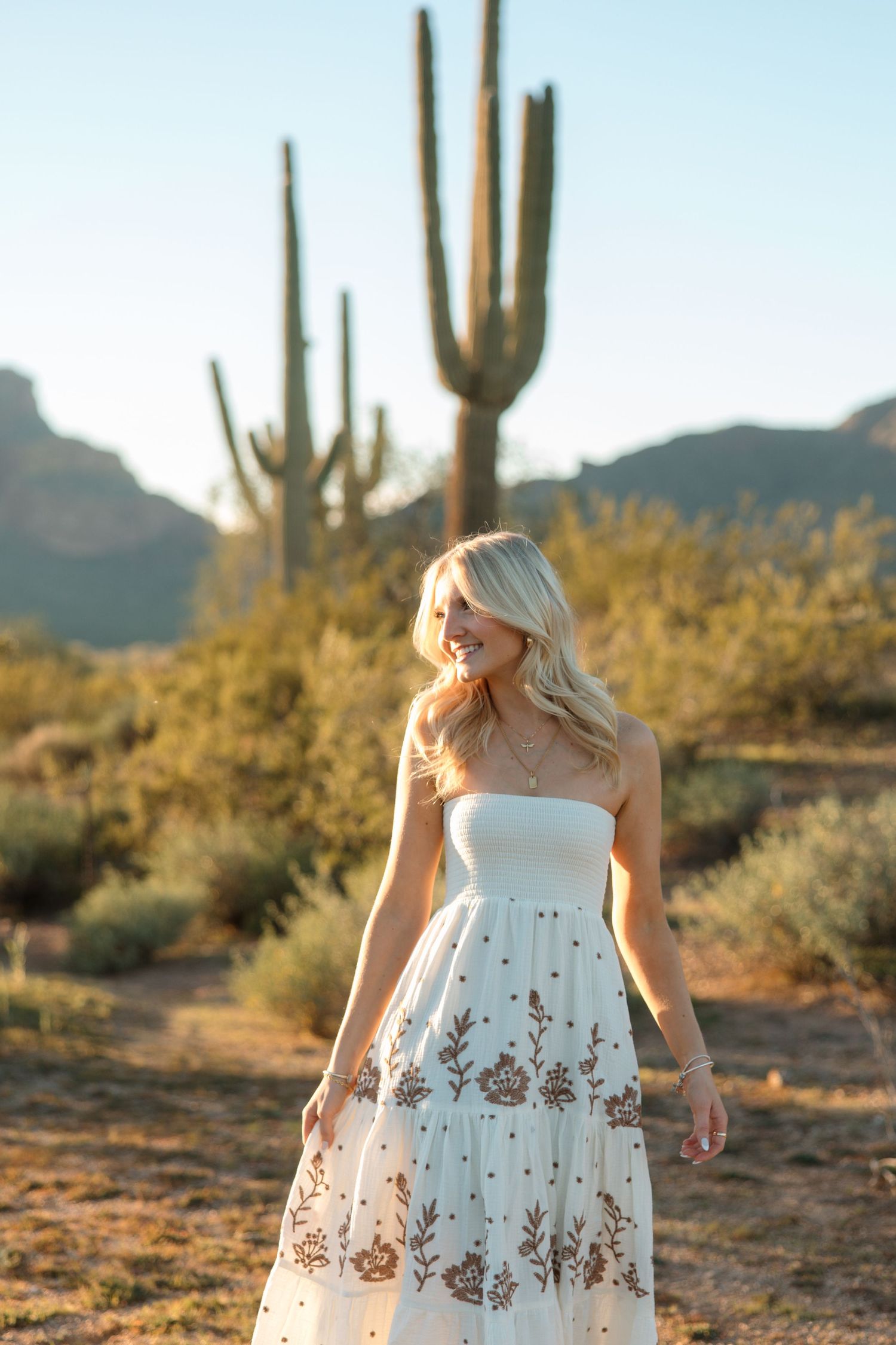 Blonde woman in white embroidered strapless dress walking through Arizona desert at sunset.
