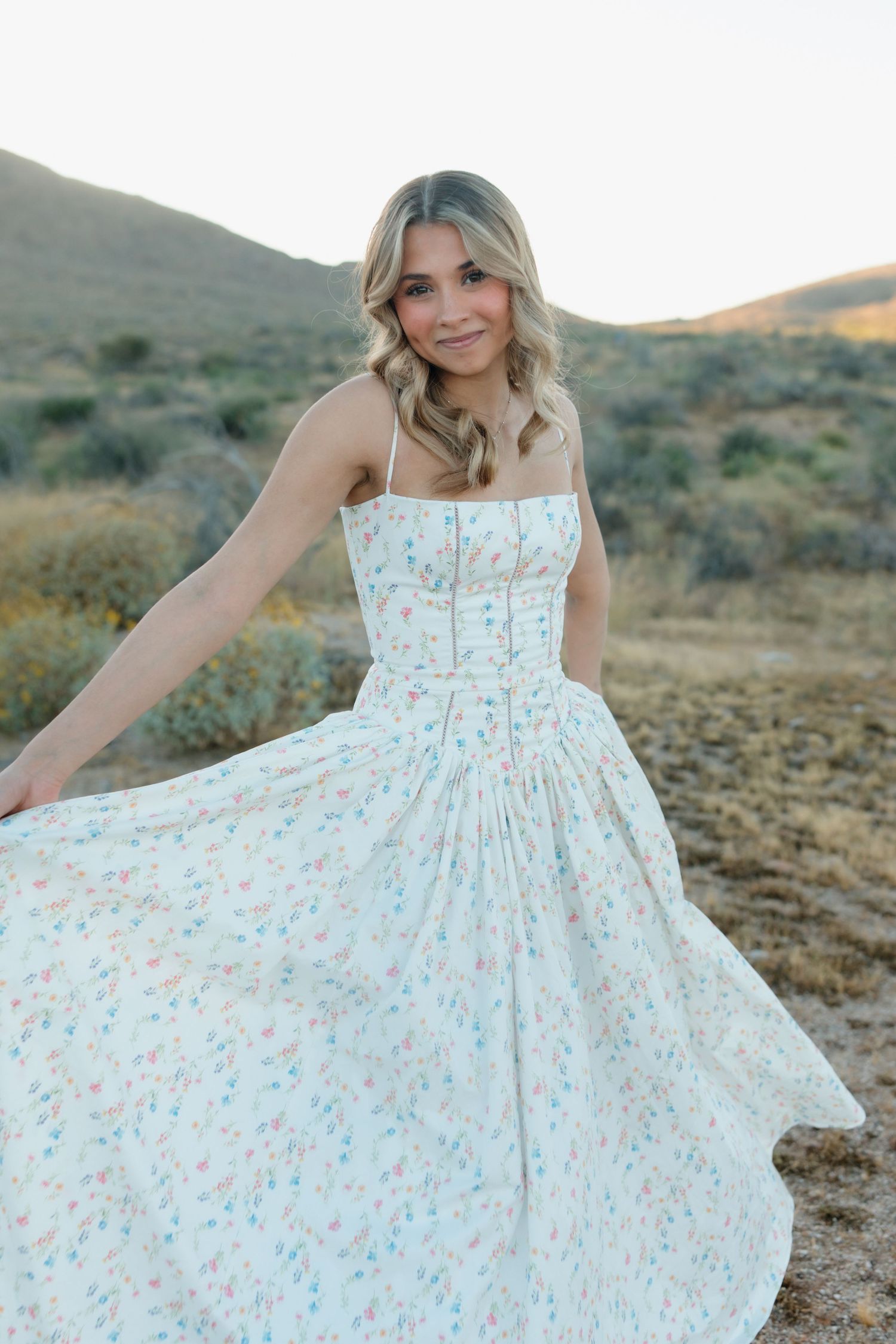 Smiling blonde woman in white floral dress twirling in desert landscape at dusk.
