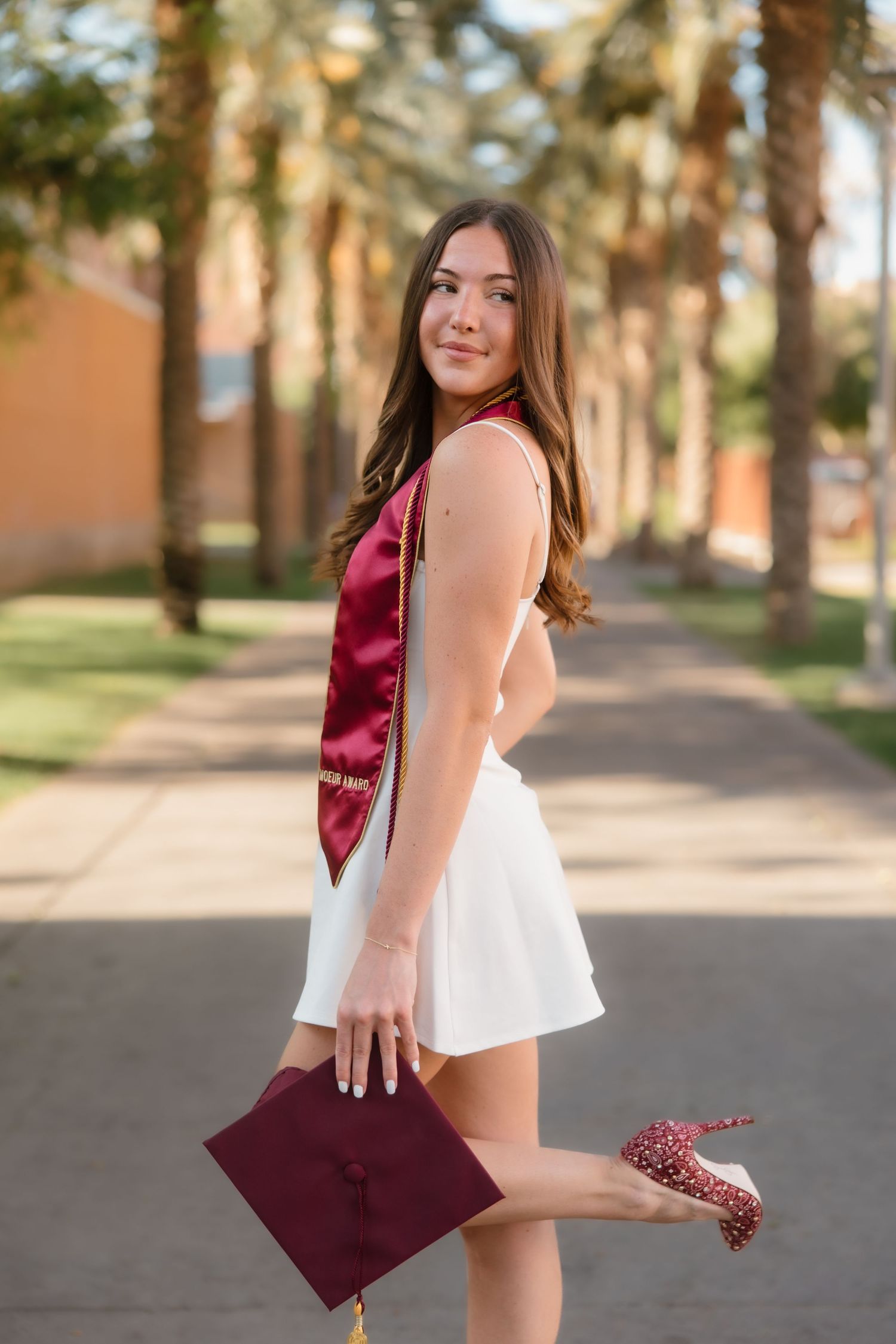 Smiling graduate in white dress holds maroon cap and stole along palm tree-lined university walkway.