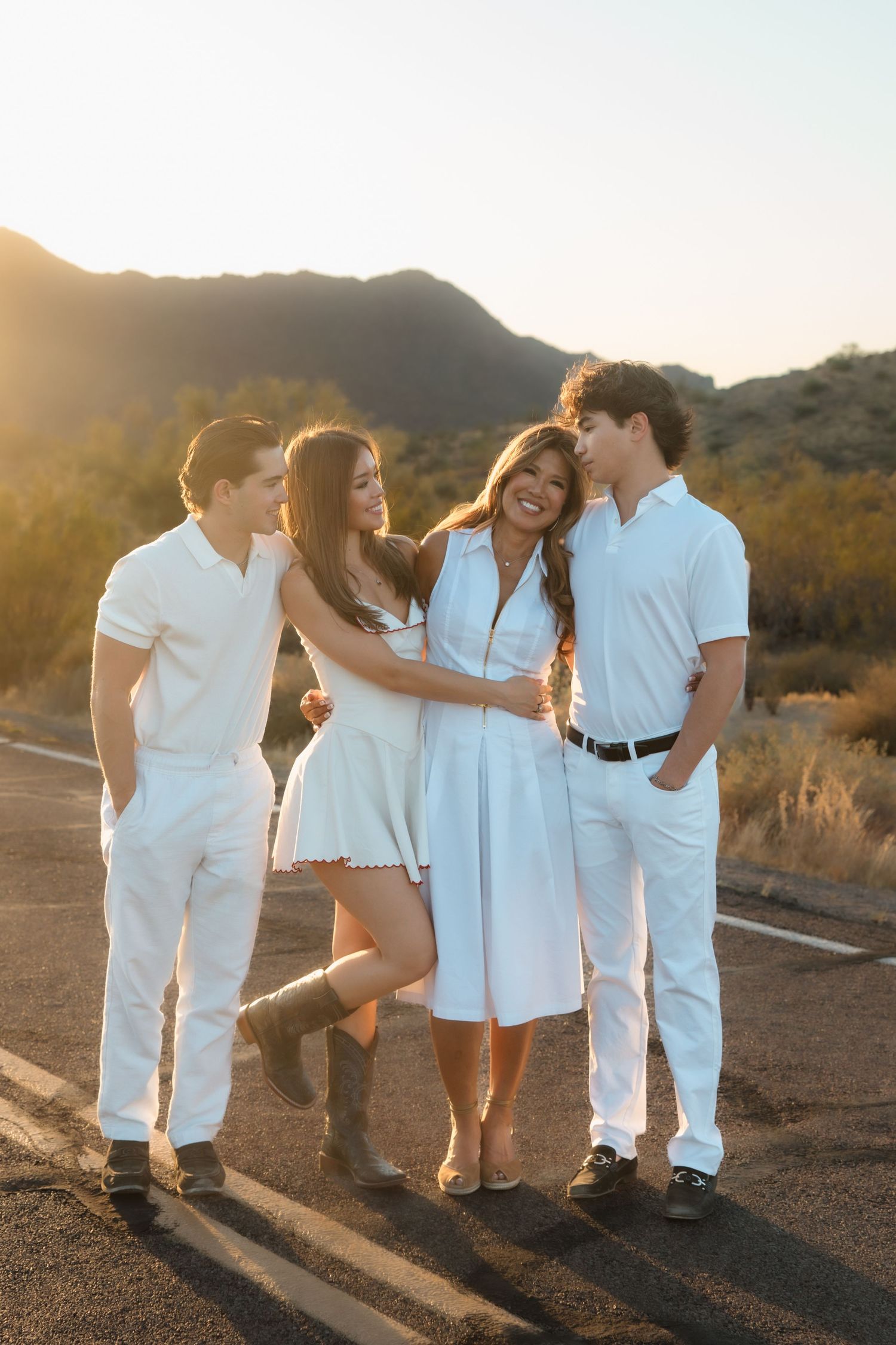 Family of four in white outfits laughing together on desert road during golden hour sunset.