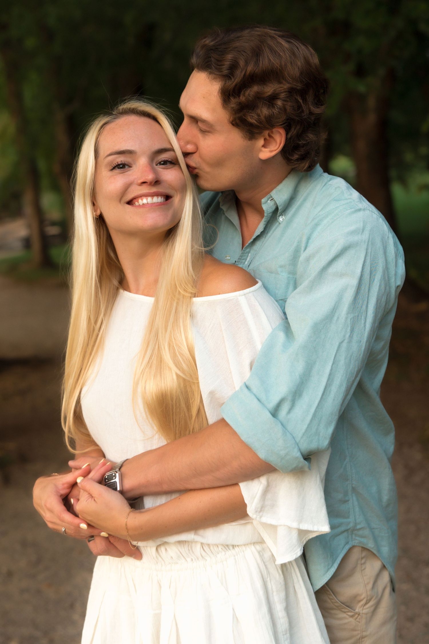 Man kissing smiling blonde woman on the cheek while embracing her from behind in a park setting.