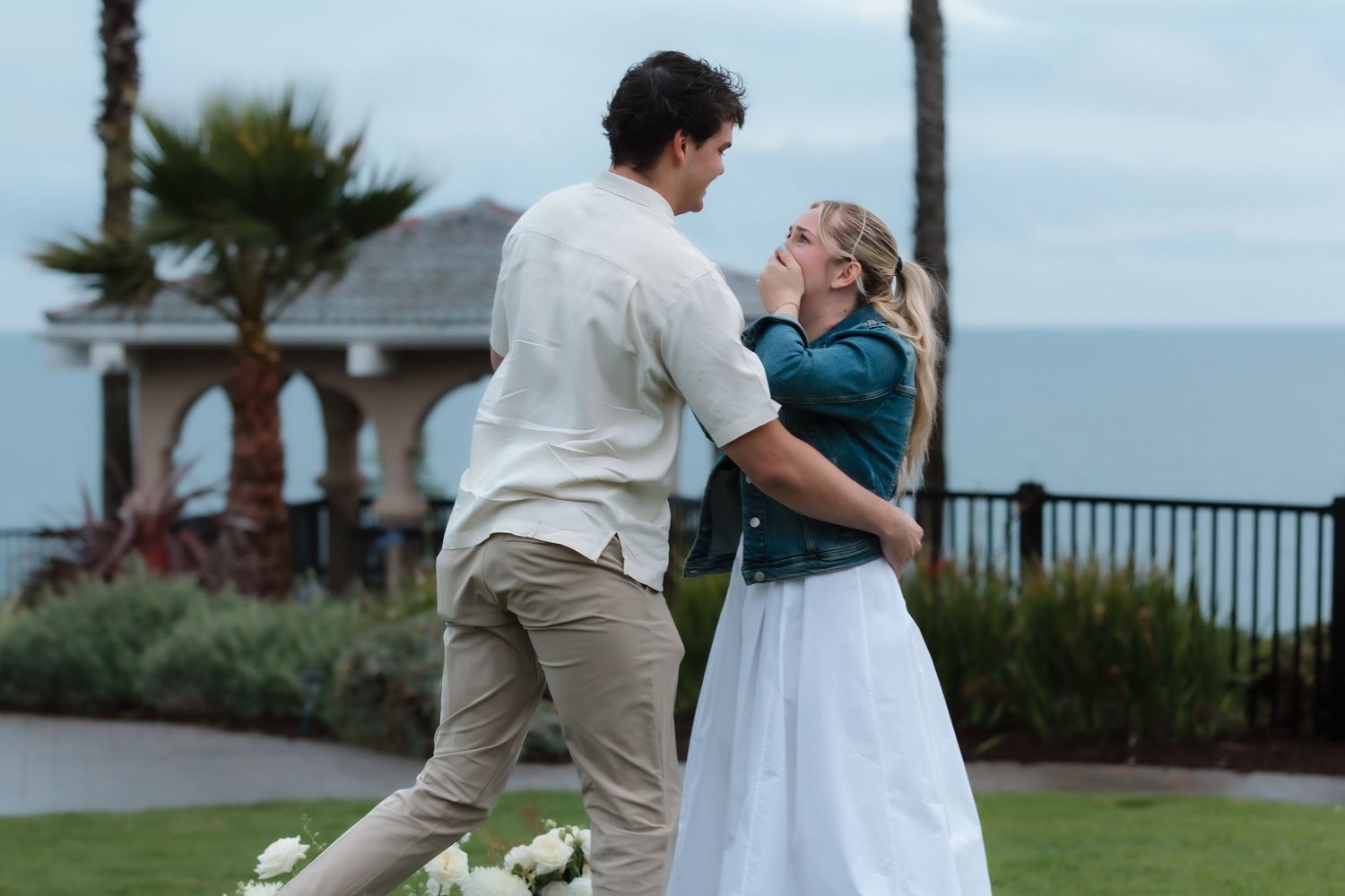 Couple dancing outdoors near ocean with palm trees and gazebo in background on overcast day.