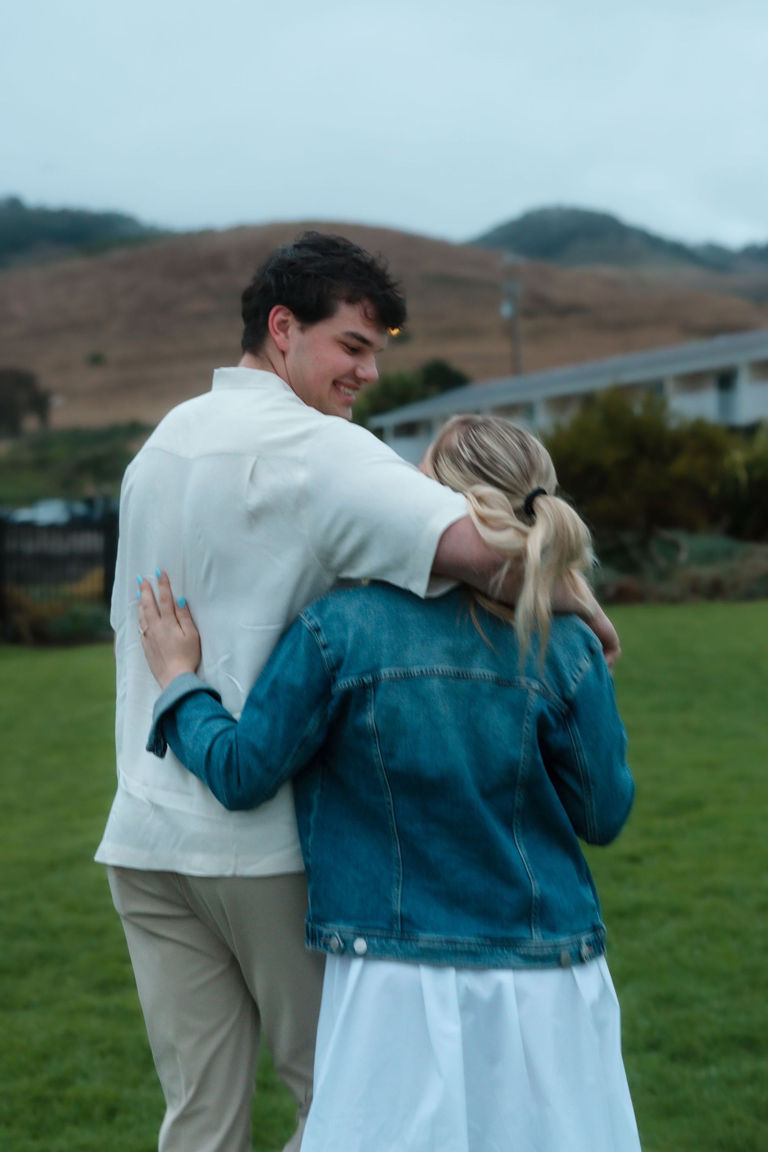 A couple embracing outdoors on green grass with misty hills in the background, man in white shirt smiling warmly.