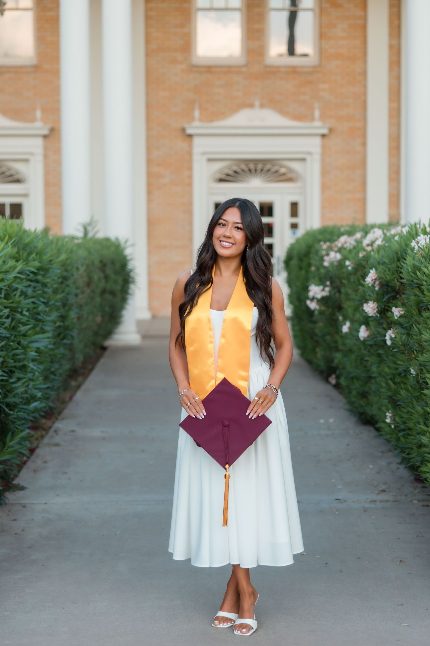 Smiling graduate in white dress holds maroon cap with gold tassel, wearing gold stole outside brick building.
