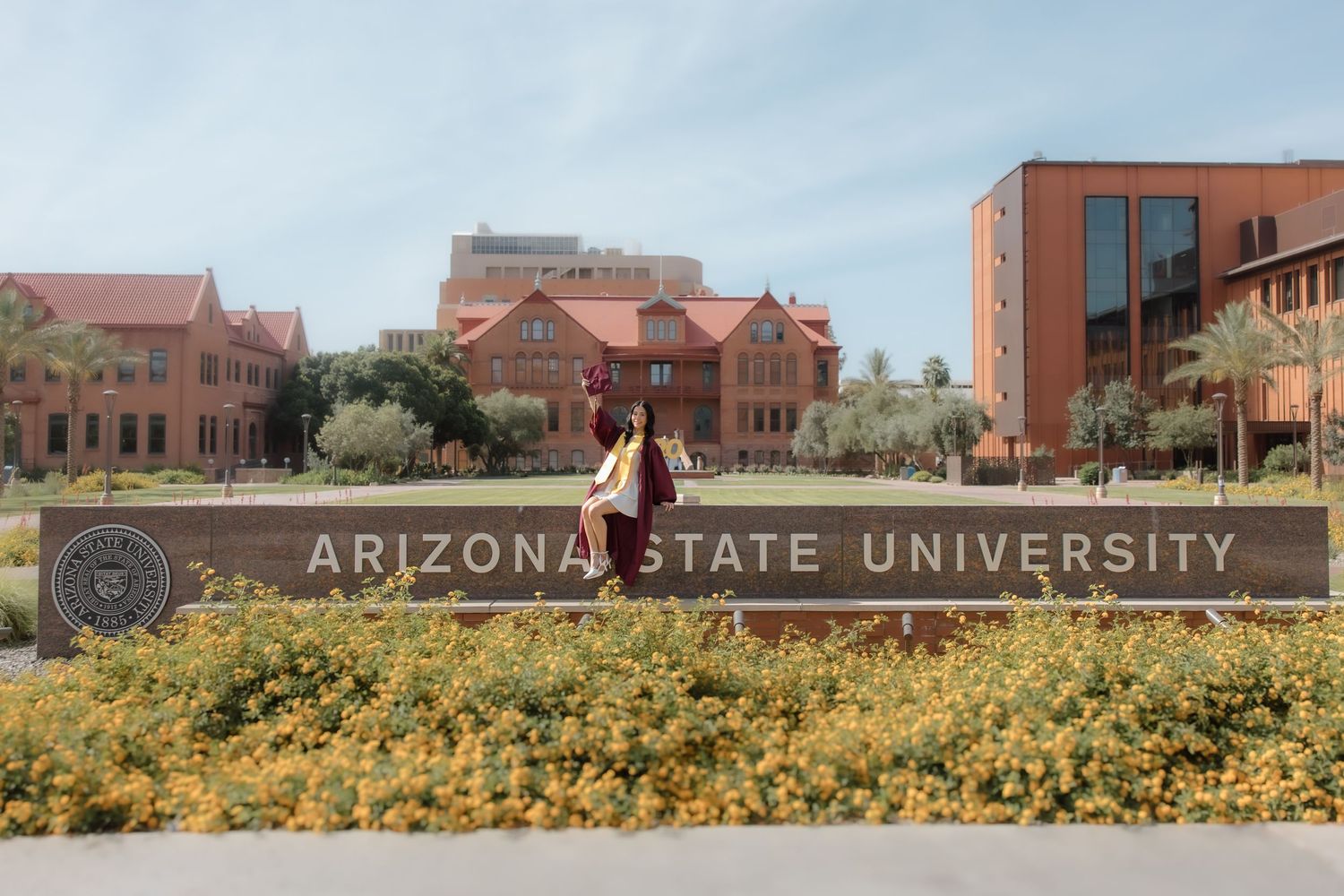 Arizona State University sign with yellow flowers, students walking on campus with red brick buildings under blue sky.