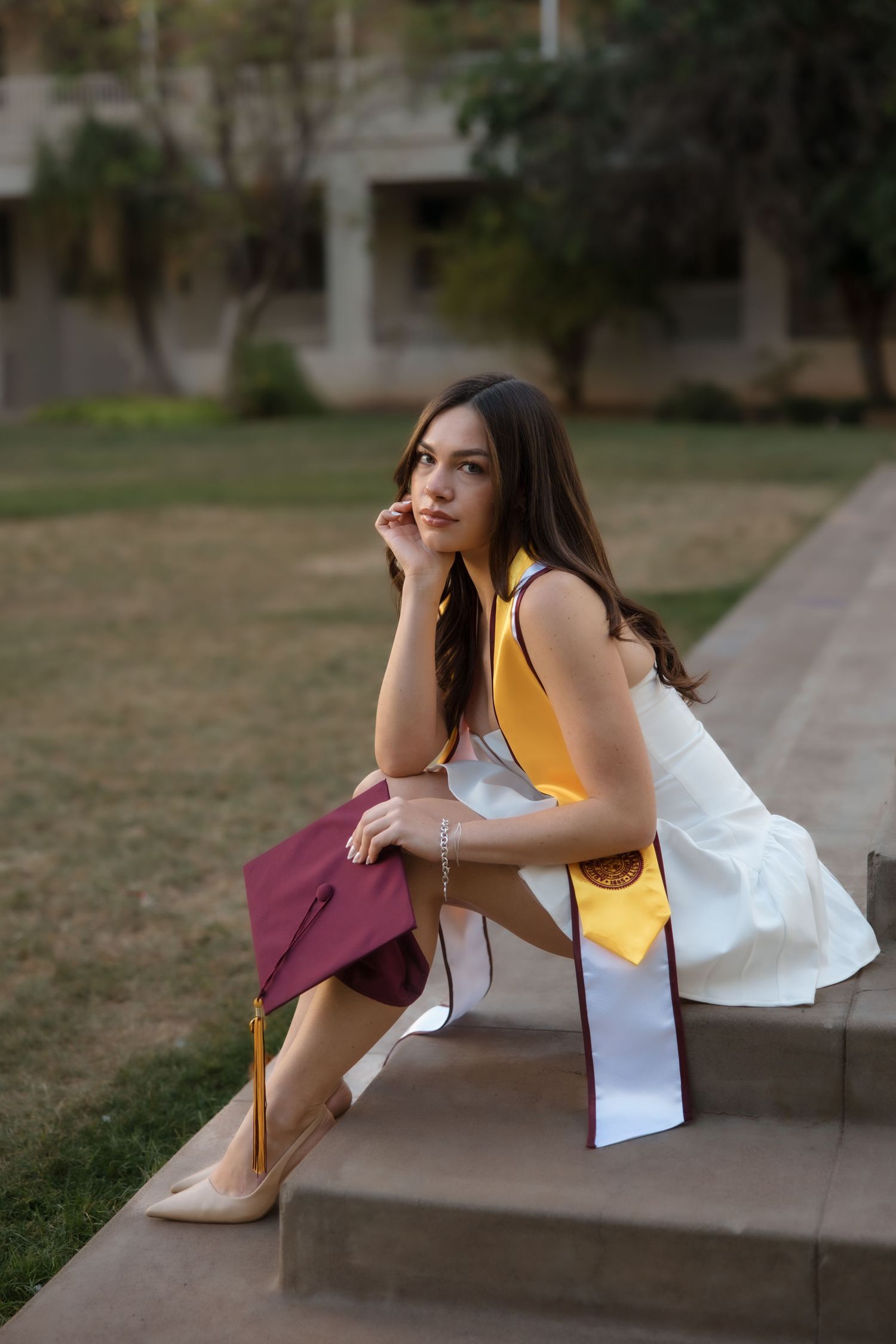 Young woman in graduation attire with maroon cap and gold stole sits on steps outdoors at university campus.
