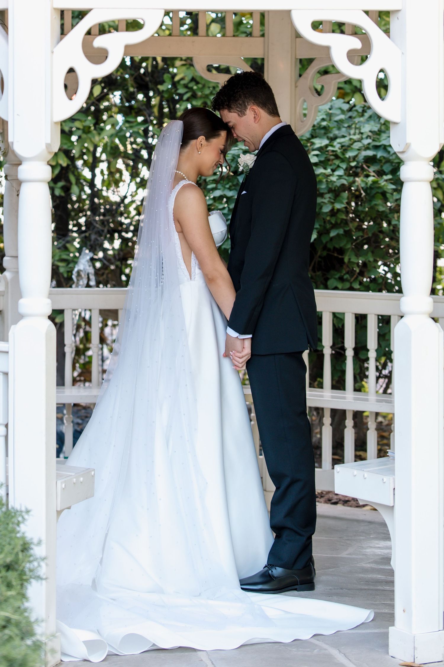Bride and groom hold hands facing each other inside a white gazebo framed by green foliage.