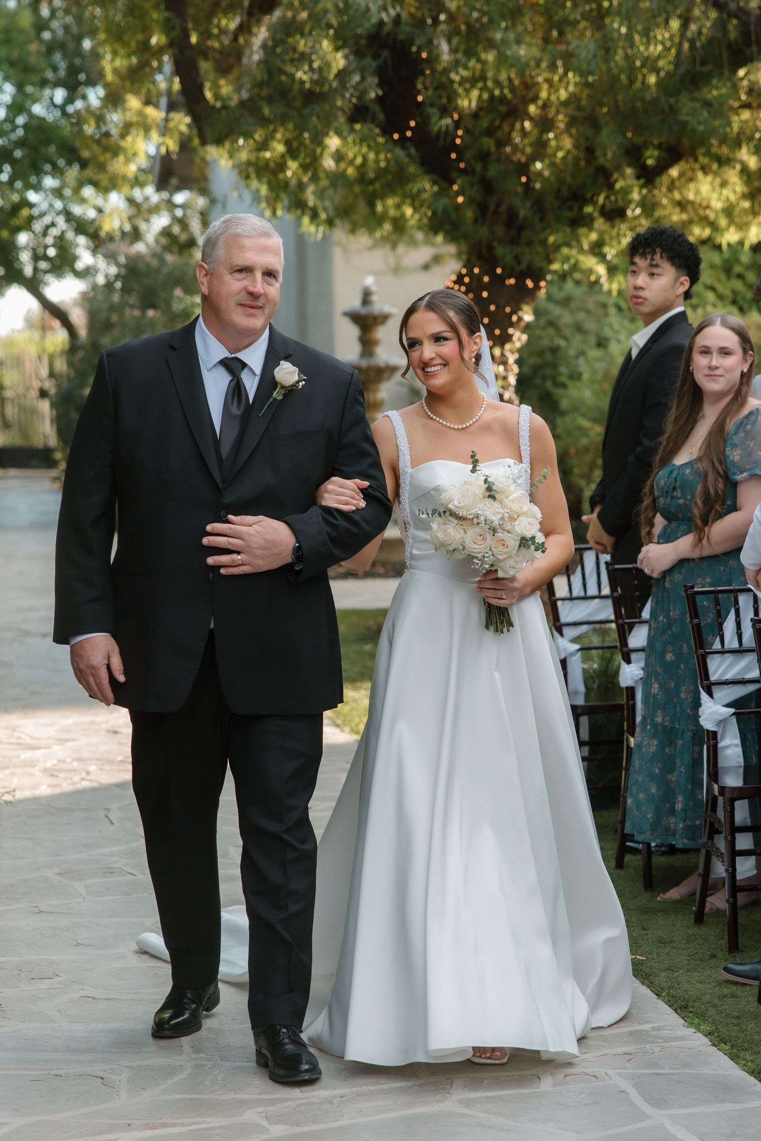 Smiling bride in white satin gown walks down outdoor aisle arm in arm with her father.
