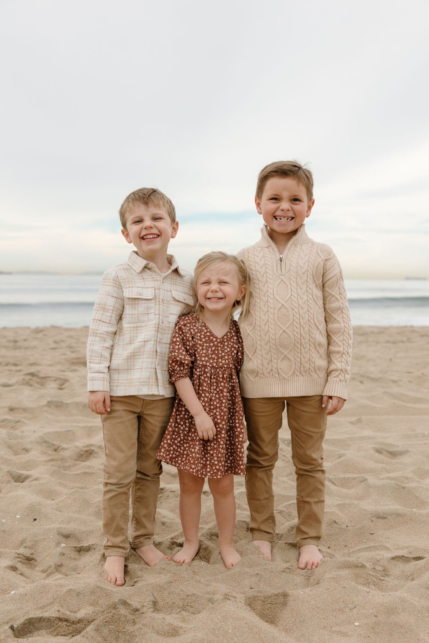 Three young siblings smiling together on a sandy beach, dressed in coordinating neutral and brown outfits.