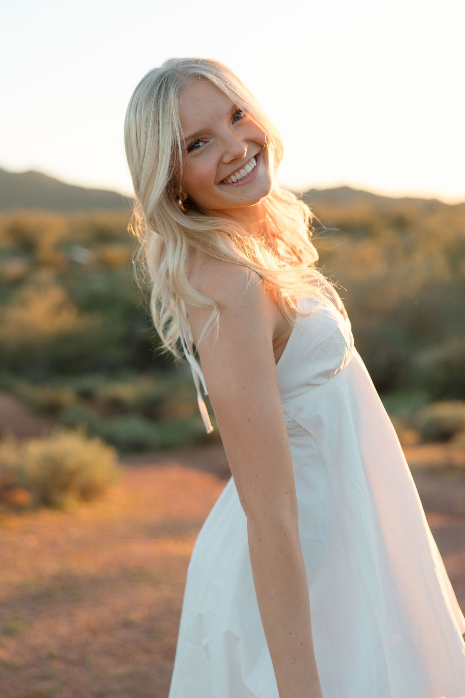 Smiling blonde woman in white strapless dress posing outdoors in desert landscape at golden hour sunset.