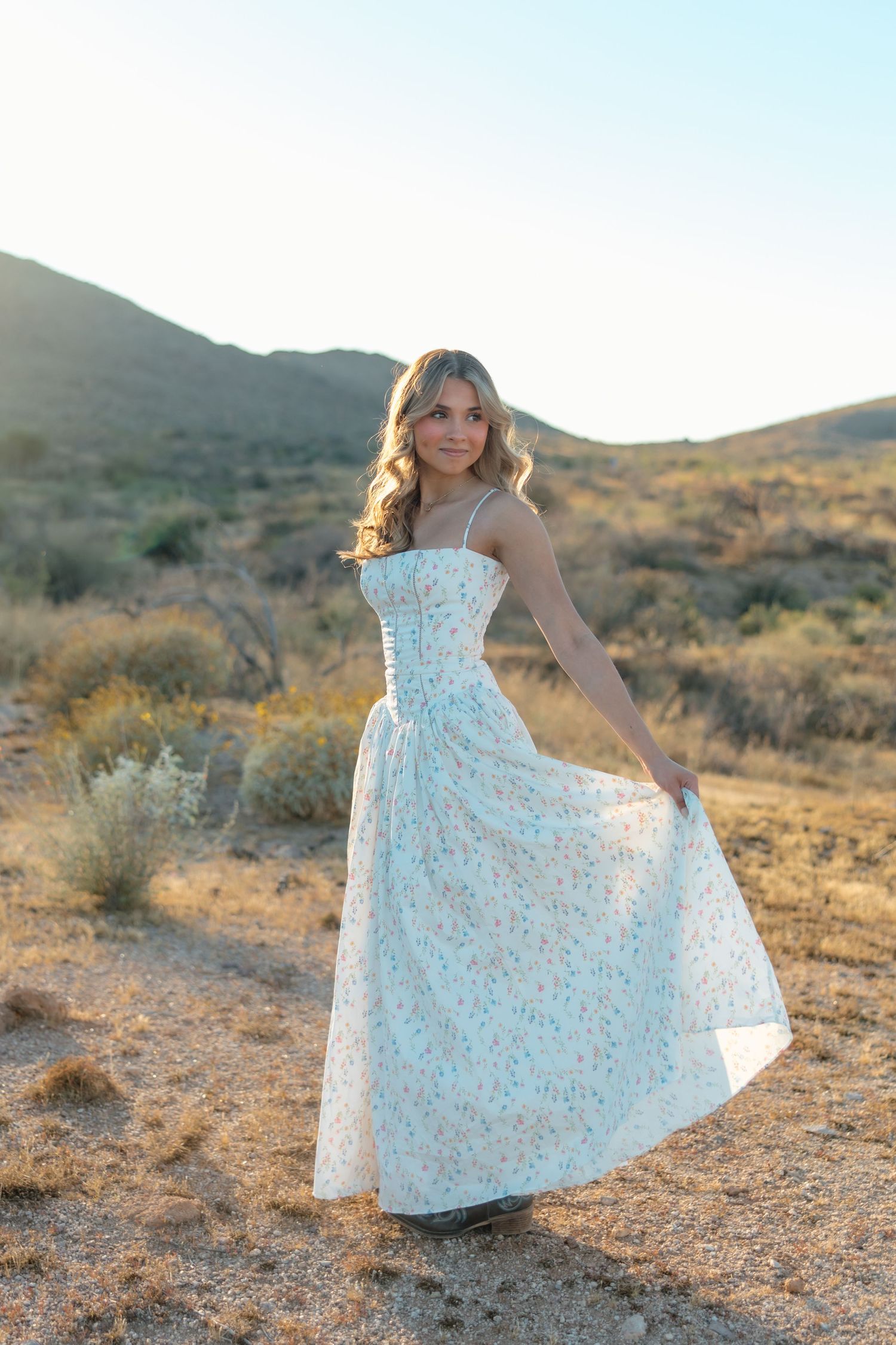 Young woman in white floral maxi dress twirling in desert landscape at golden hour with mountains in background.