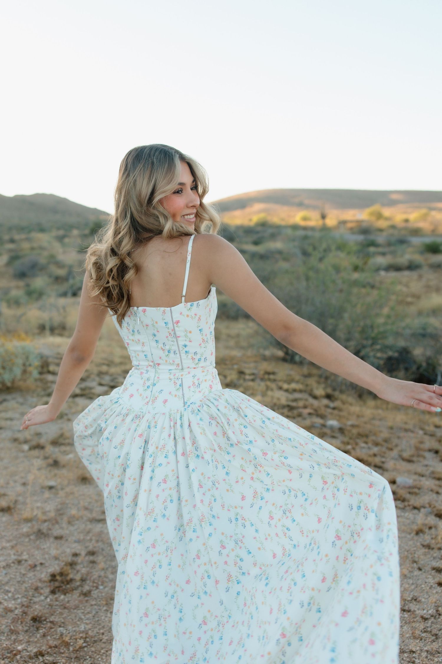 A woman in a floral sundress twirls outdoors in a desert landscape, smiling over her shoulder at golden hour.