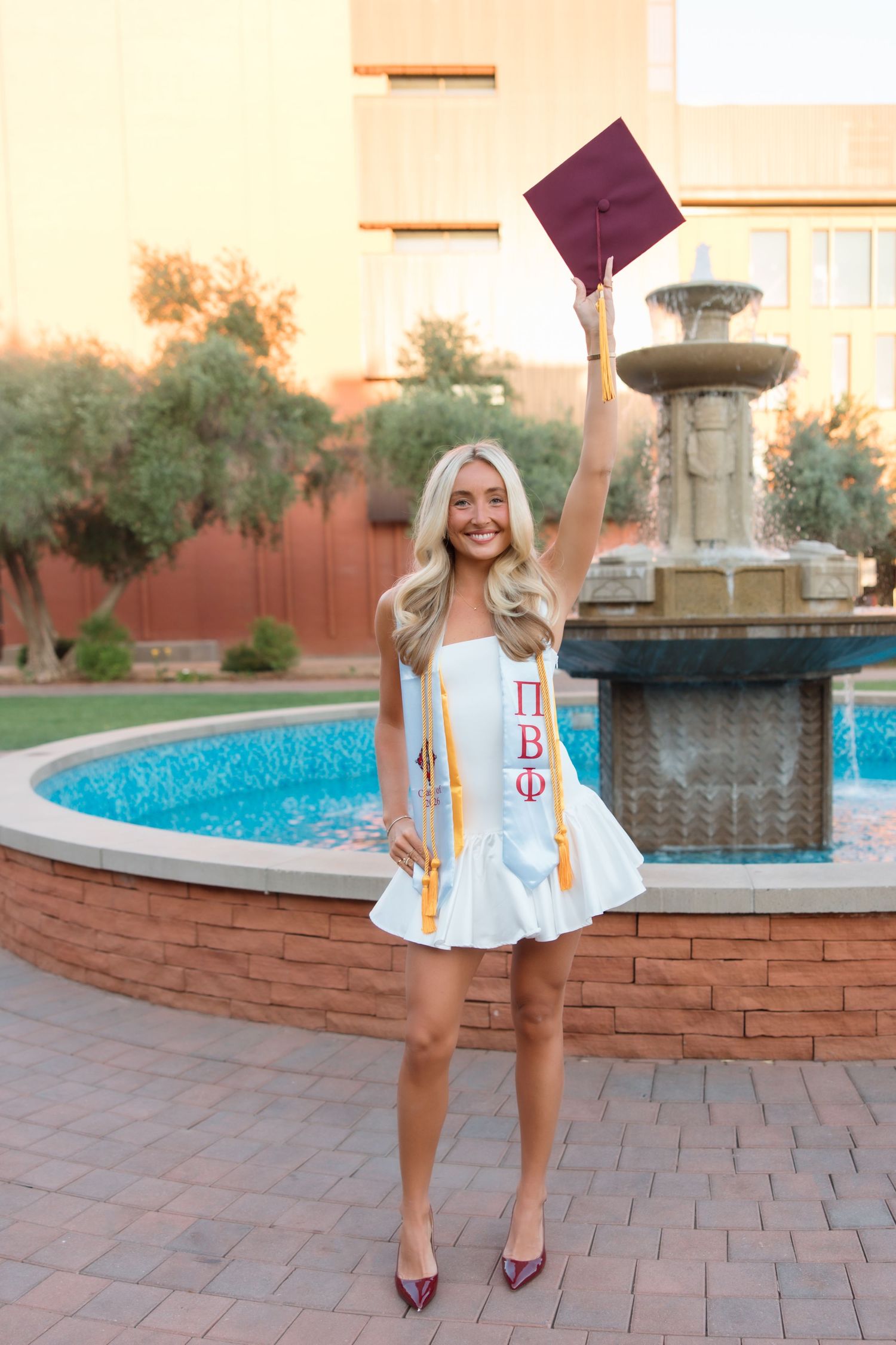 Happy blonde graduate in white dress raises maroon cap by fountain wearing Pi Beta Phi sash and maroon heels.
