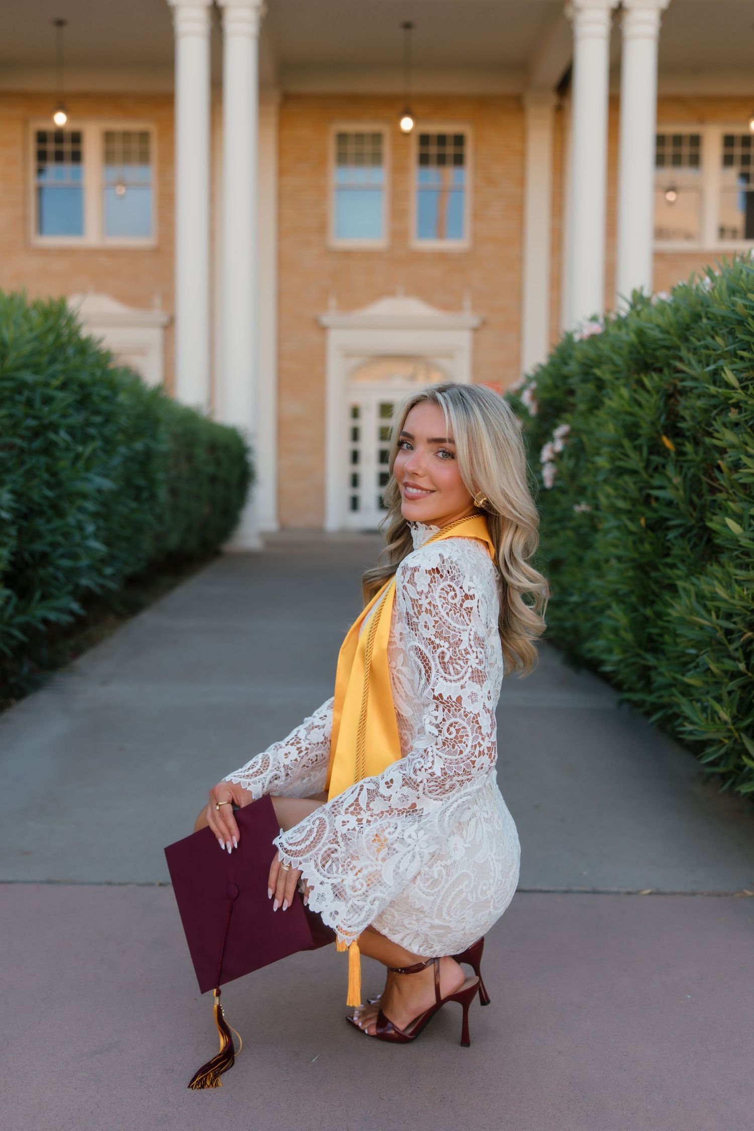 Blonde woman in white lace dress kneels with maroon graduation cap outside brick building.