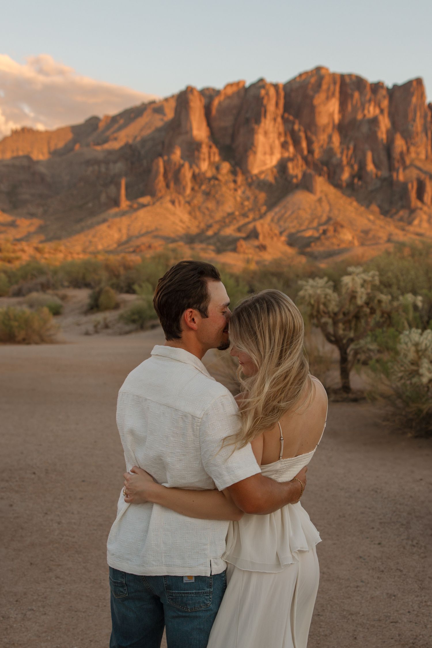 Couple embracing and kissing at sunset with Superstition Mountains glowing orange in Arizona desert.