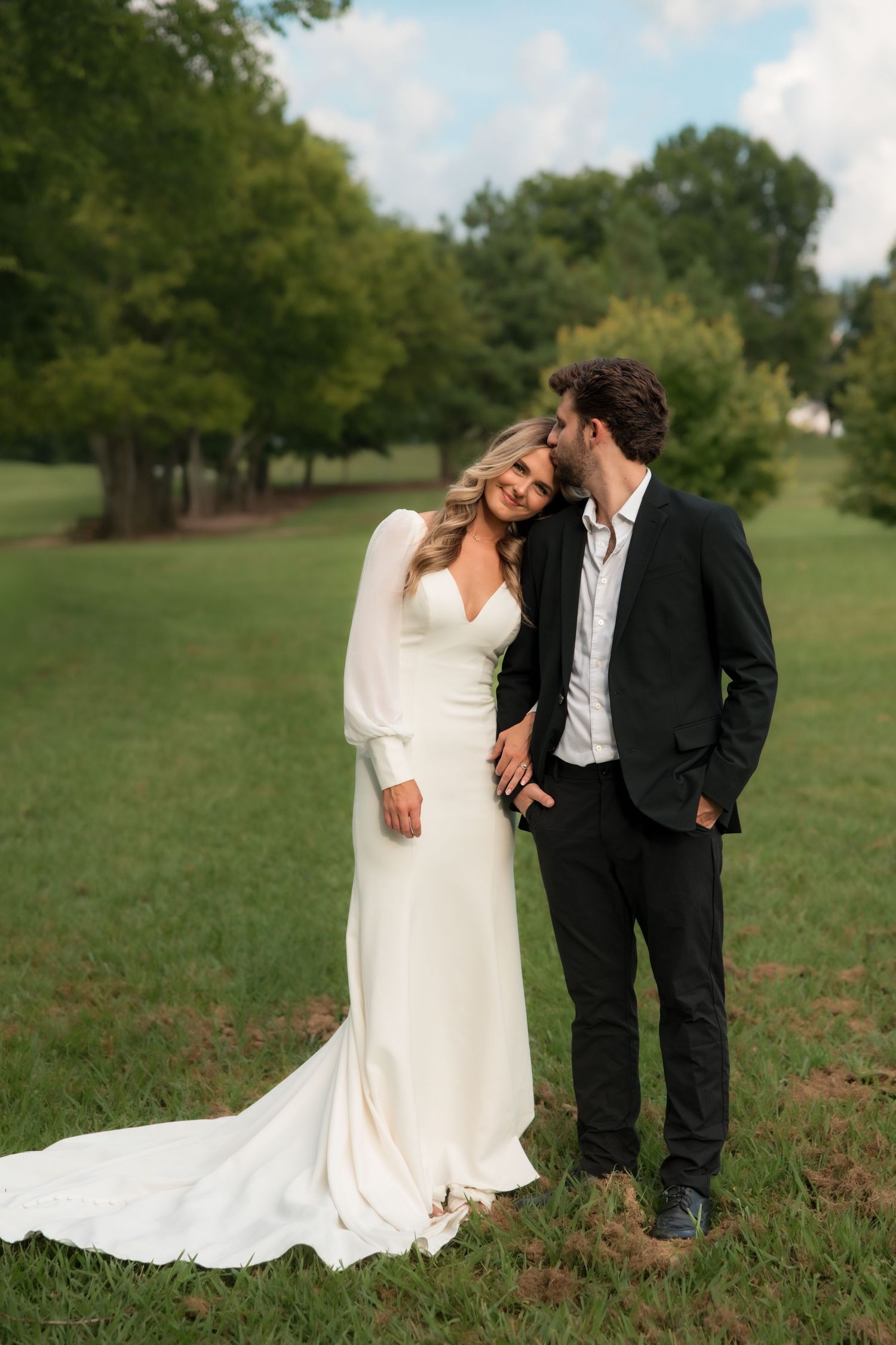 Groom in black suit kissing bride on cheek as she smiles, standing in green field with trees behind them.