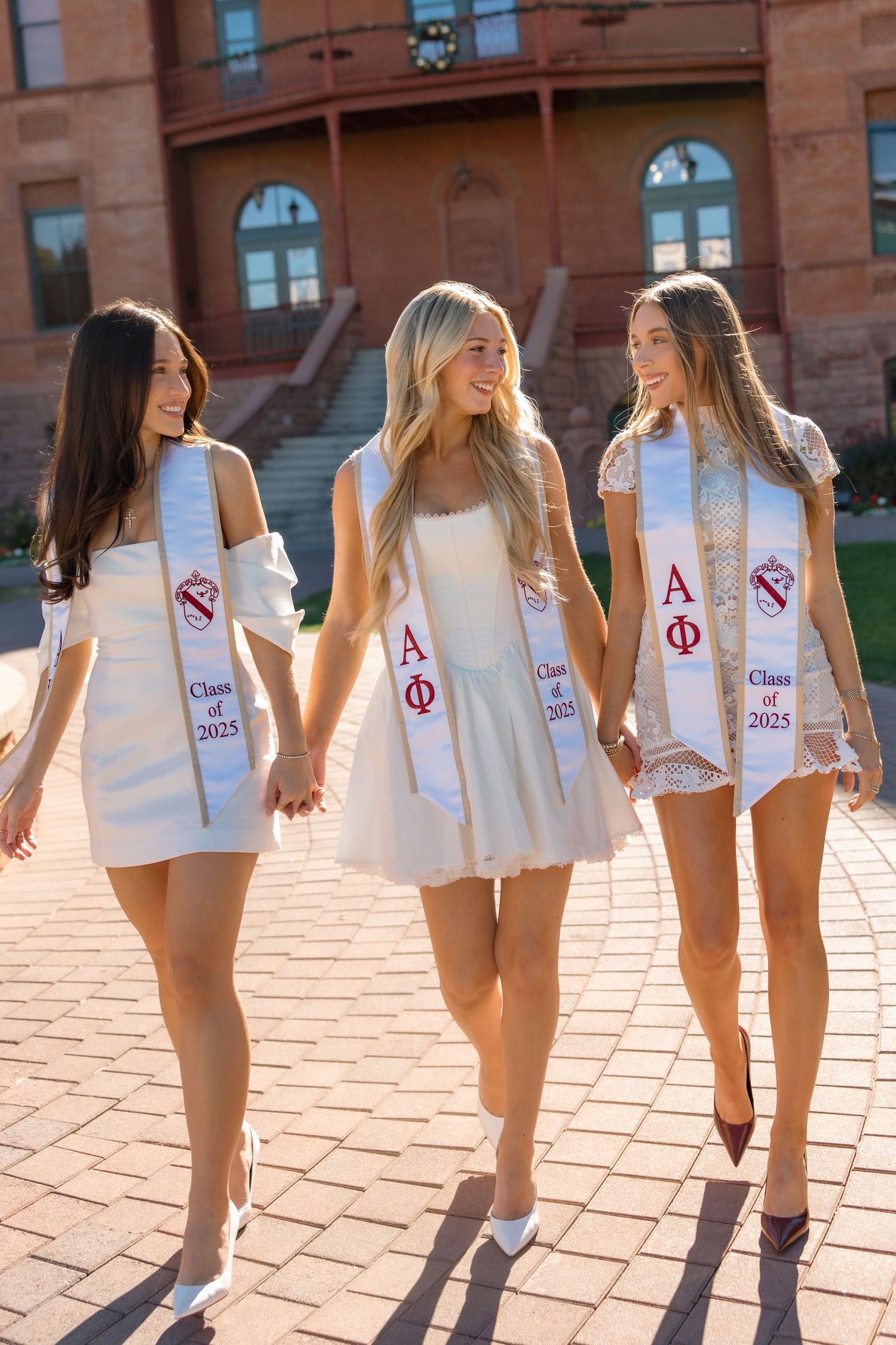 Three Alpha Phi sorority graduates in white dresses walk hand-in-hand wearing Class of 2025 stoles.