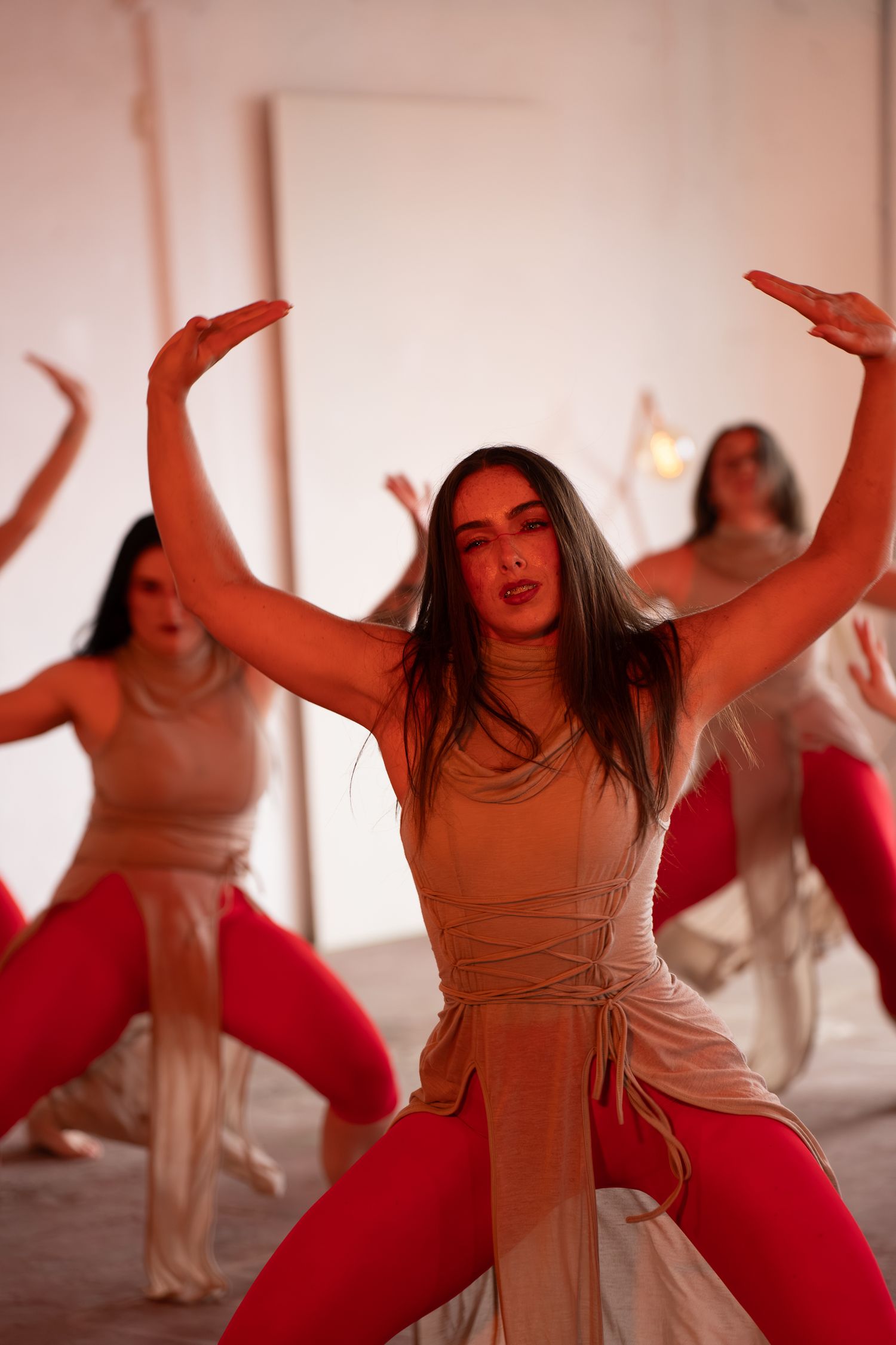 Dancers in red leggings and beige wrap tops perform a synchronized routine, with arms raised, in a softly lit studio.