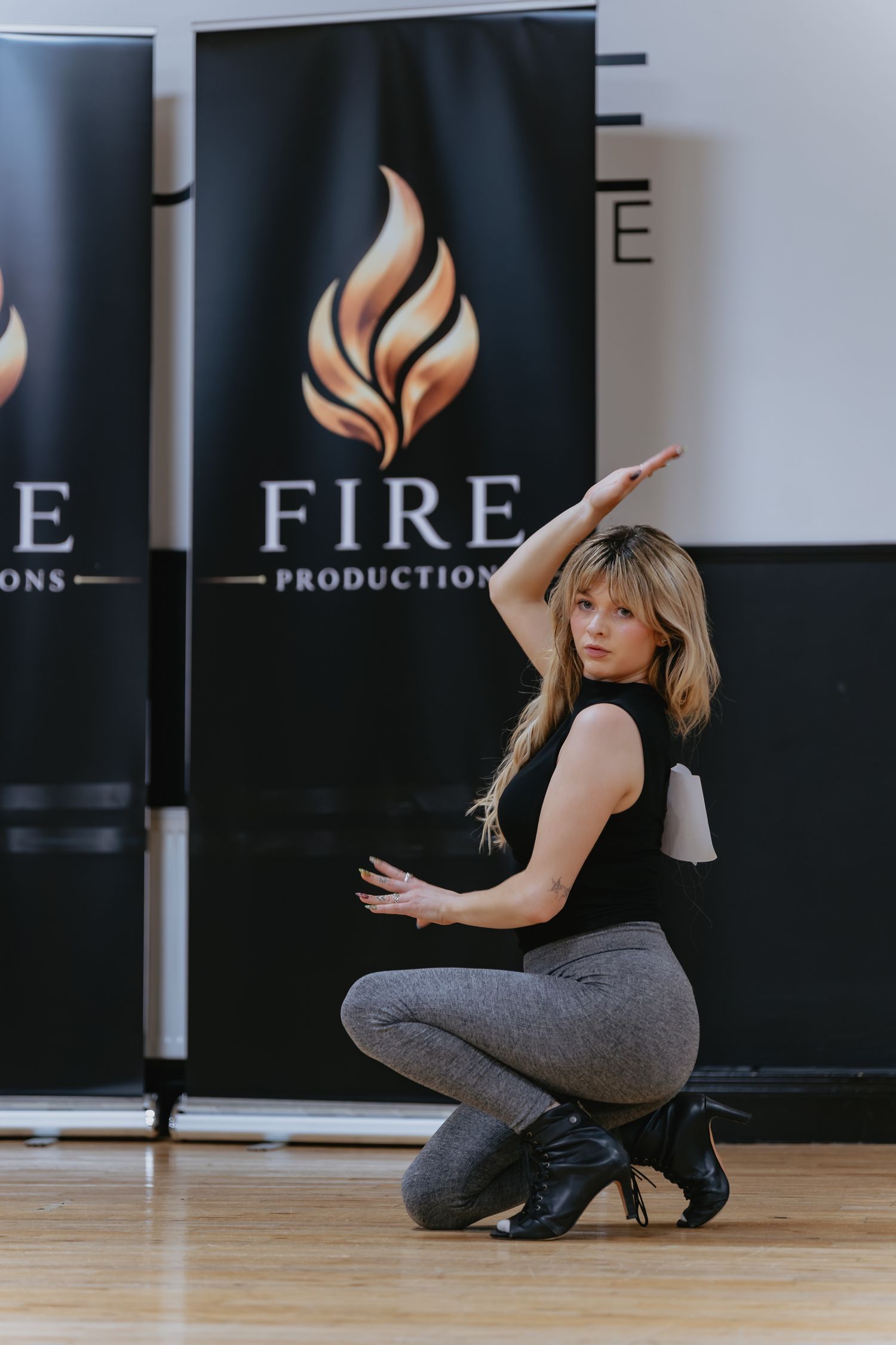 Dancer with long blonde hair crouches in a dynamic pose on a wood floor in front of black Fire Productions banners.