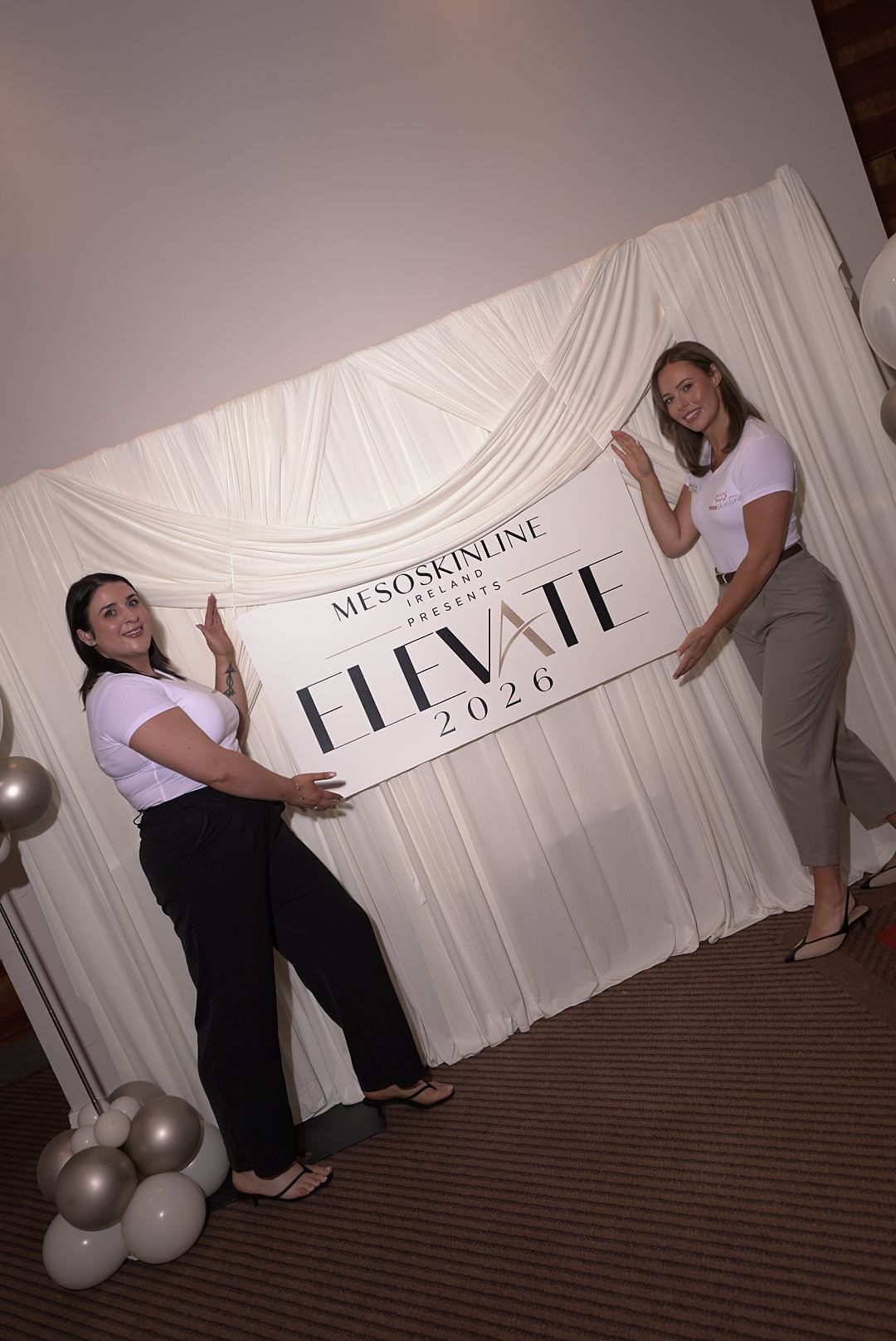 Two women in white tops pose beside a Mesoskinline Elevate 2026 sign hung on a white draped backdrop with balloons.