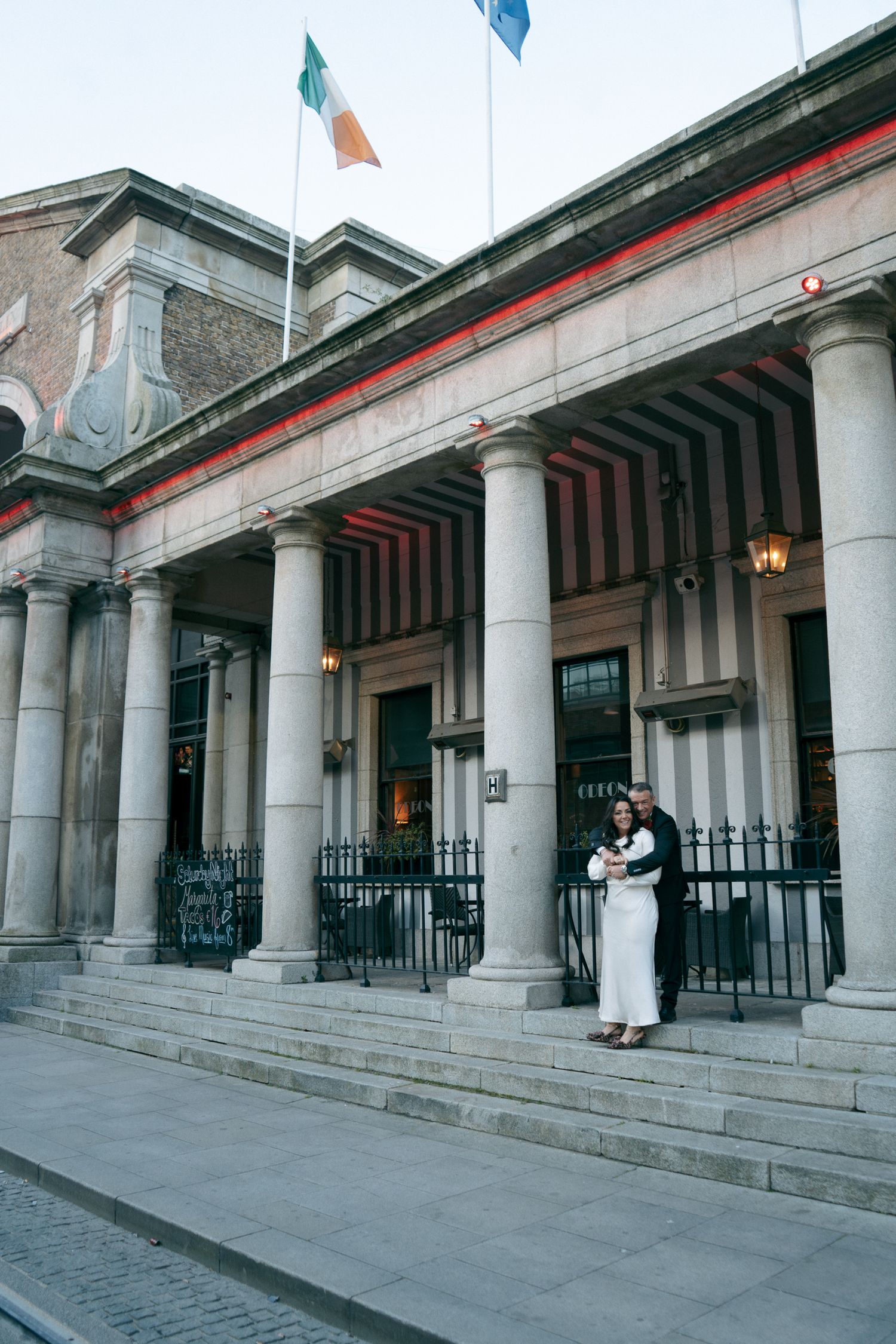 Bride and groom embrace on stone steps in front of a columned building with an Irish flag flying above, elopement photo in Dublin.